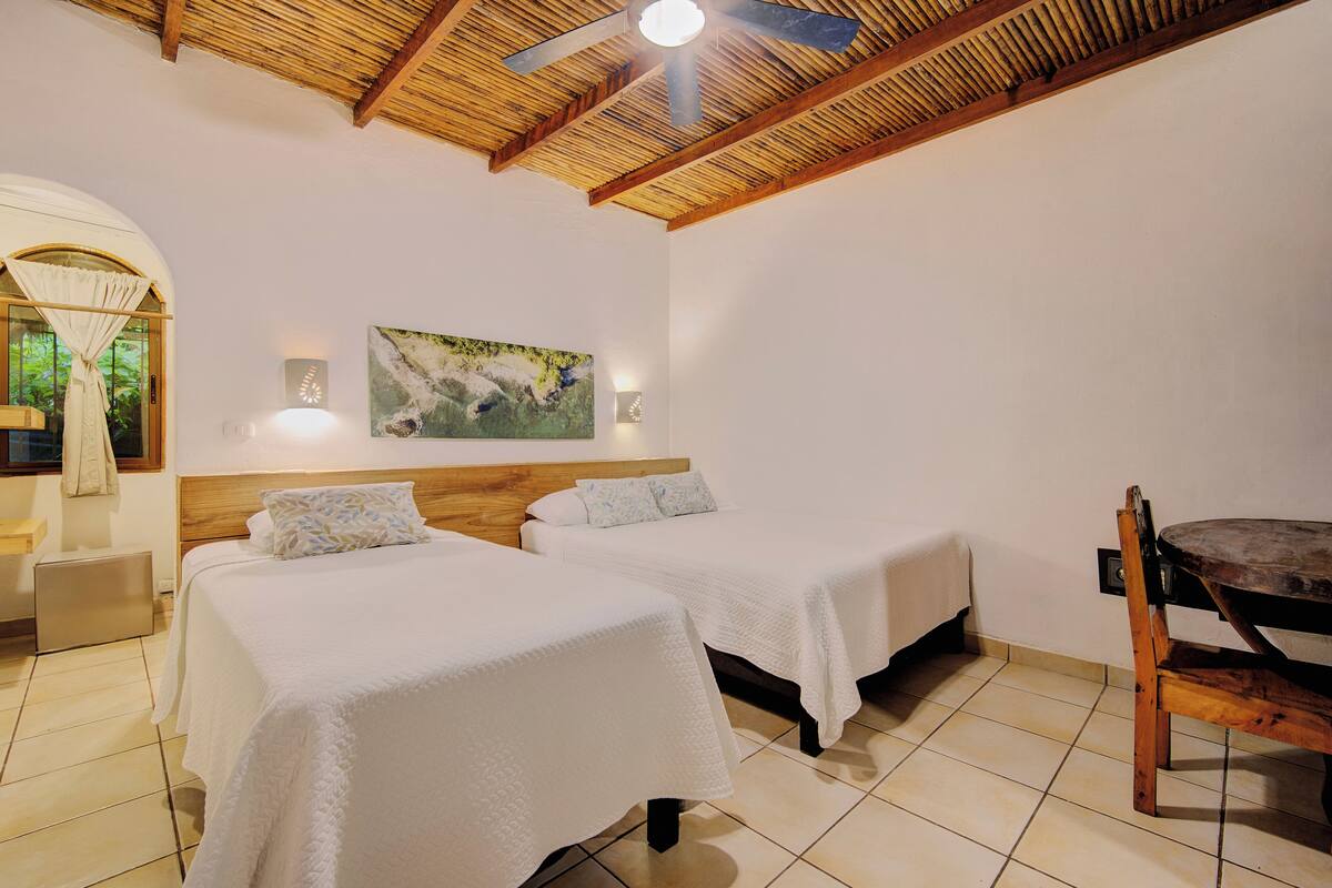 A room with two beds, a wooden ceiling, a painting on the wall, and a dining table with chairs.