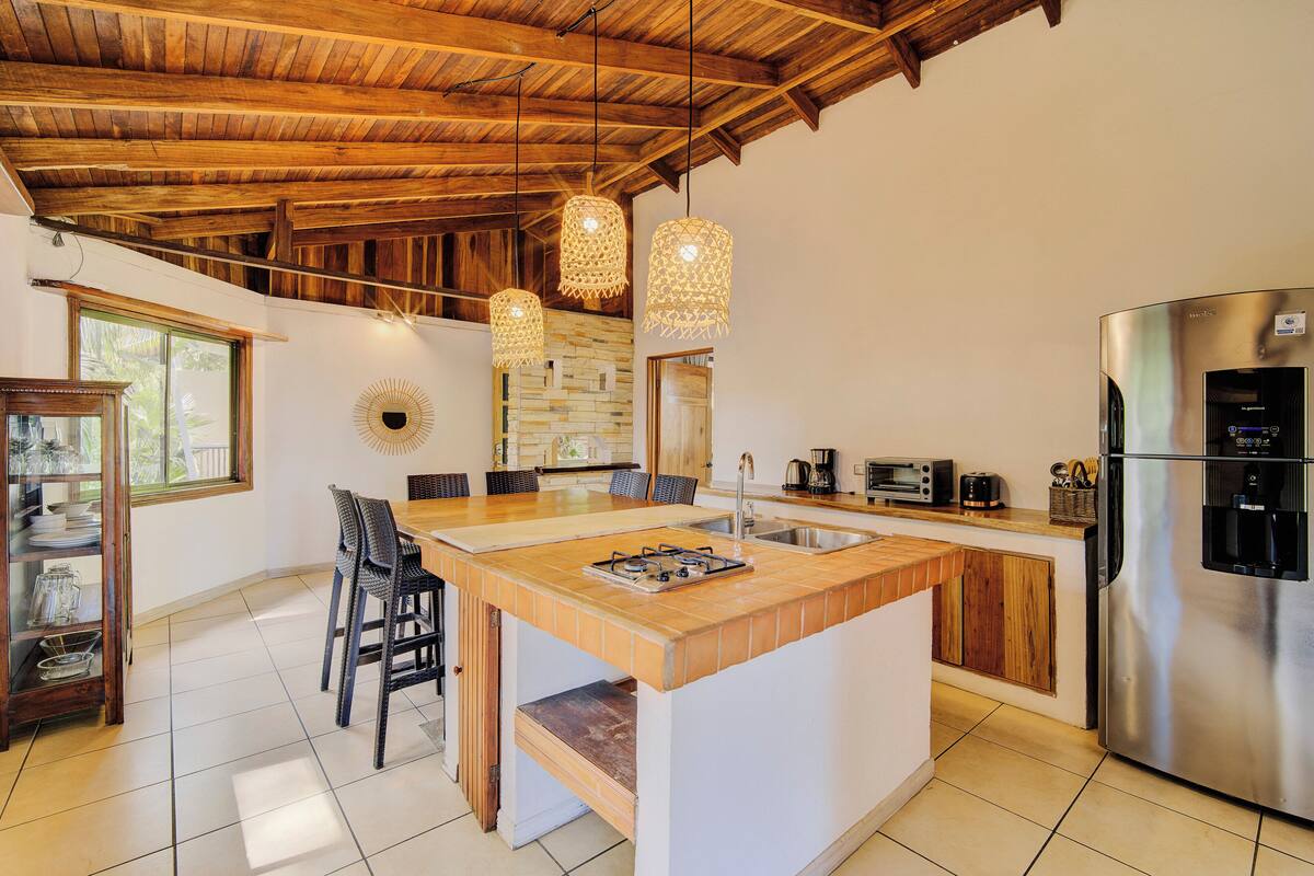 A kitchen with a central island, wooden ceiling, hanging pendant lights, a stainless steel refrigerator, and a dining area with chairs.