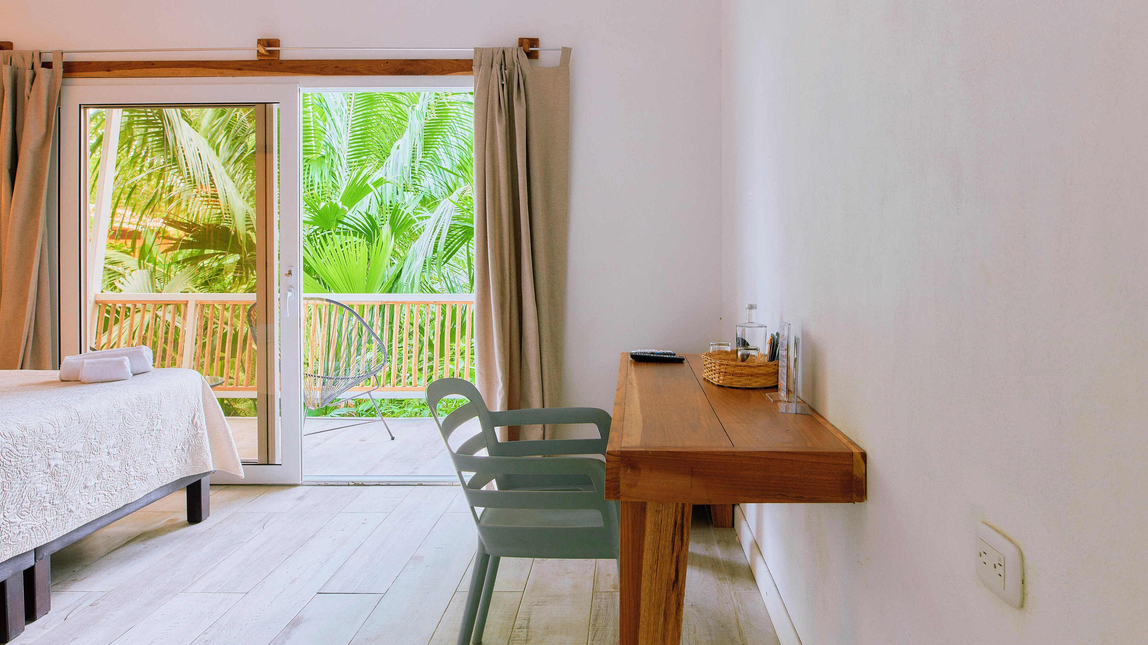 Una habitación con una cama, una mesa de madera con una cesta, una silla y vistas a la exuberante vegetación a través de una puerta corredera.