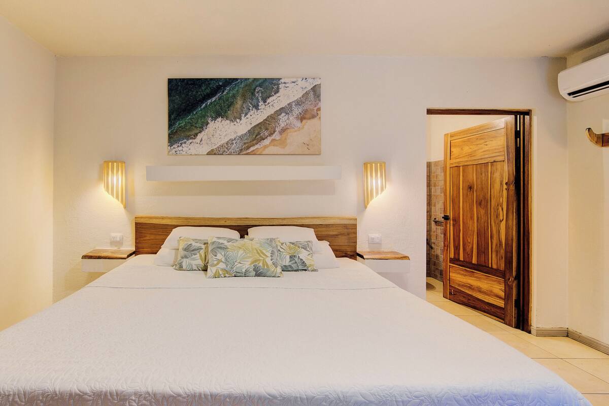 A bedroom with a large bed, two bedside tables, a wooden door, and a wall-mounted artwork.