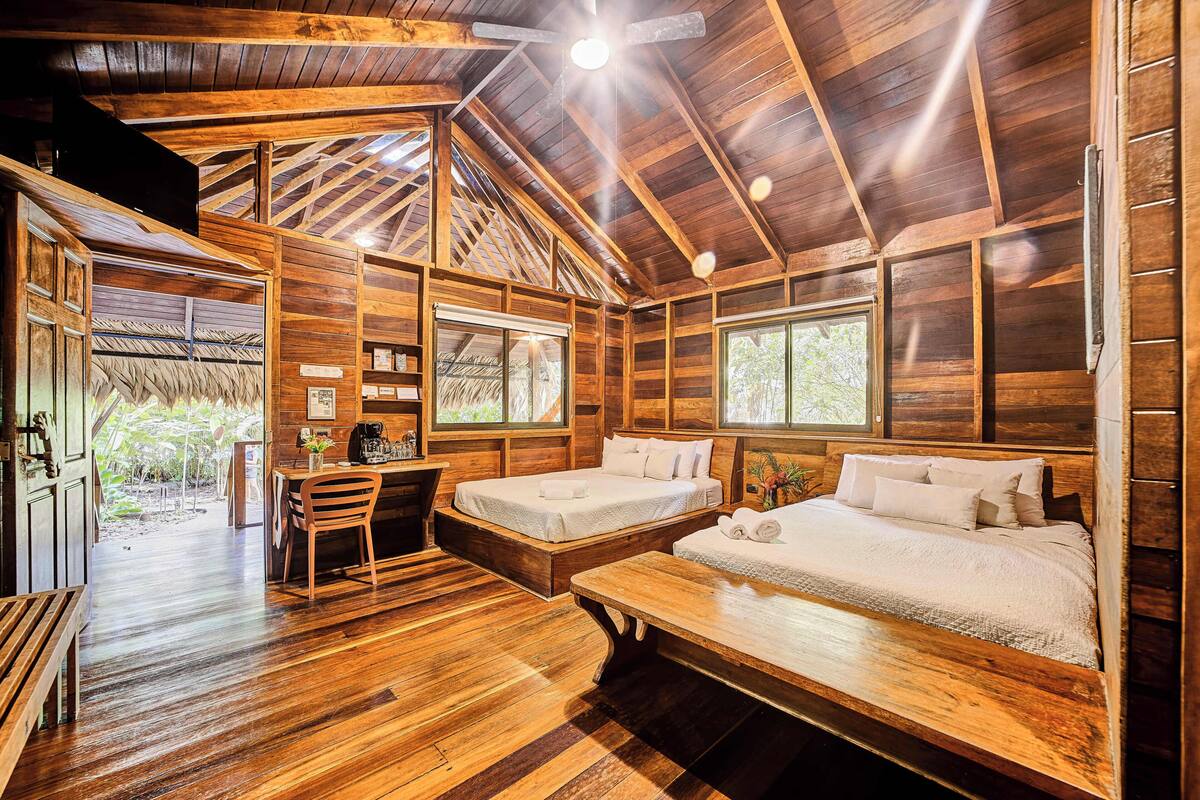 A wooden cabin interior with two beds, a dining area, and a view of the outdoors.
