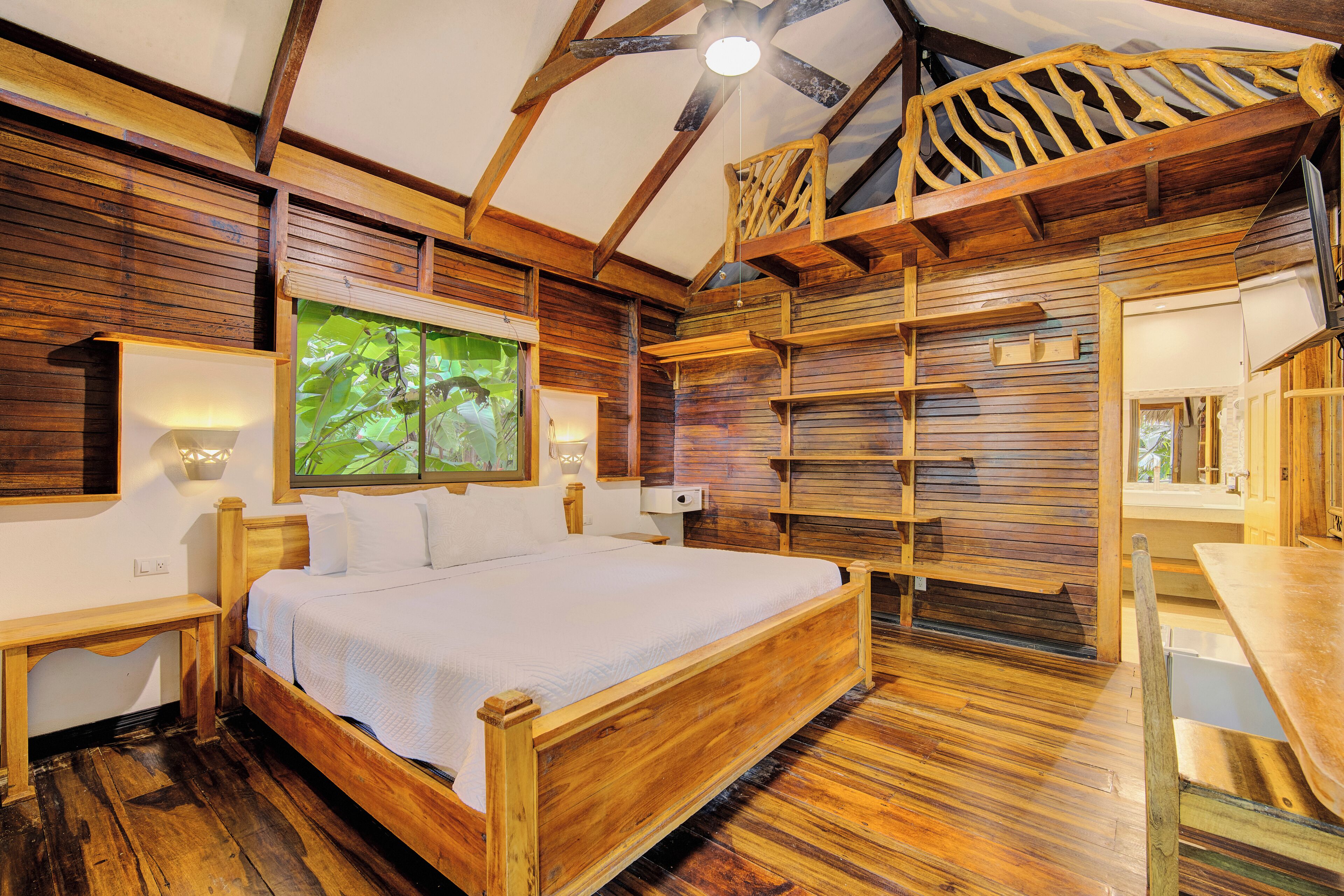 A wooden bedroom with a bed, bedside tables, a window with a view, and a ceiling fan.