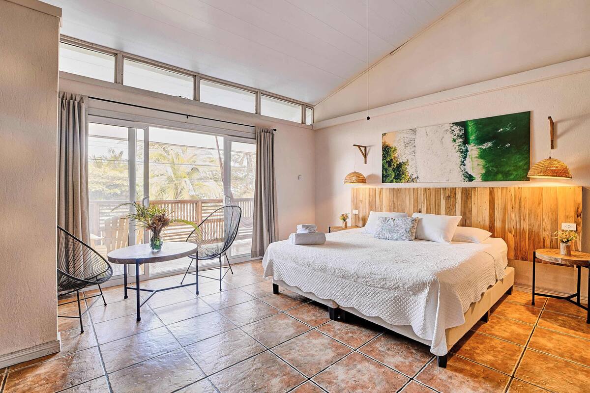 A bedroom with a large bed, a balcony with a chair, a small table with a vase, and a painting on the wall.