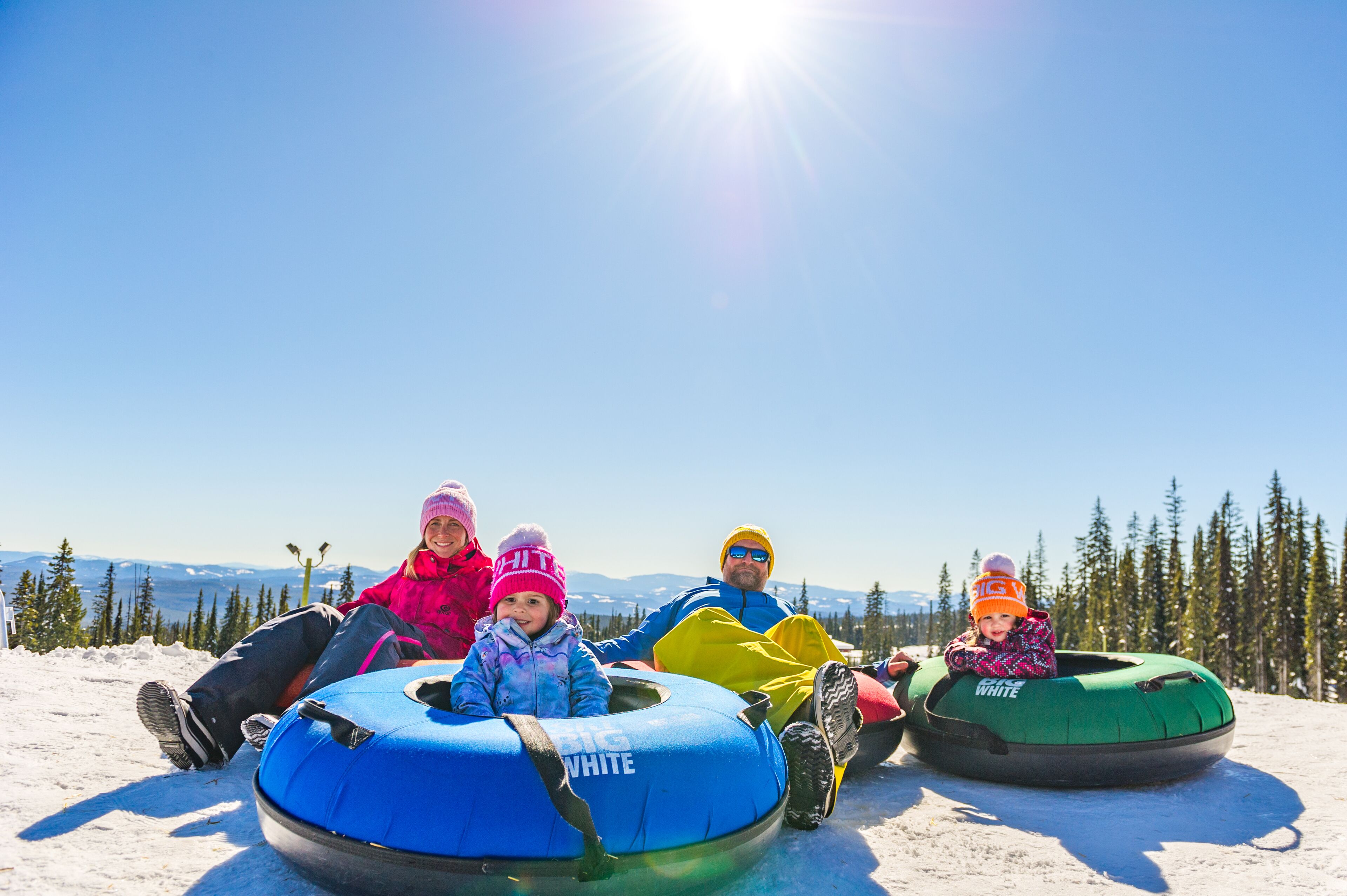 A group of people sitting on snow tubes on a snowy slope with a clear sky and trees in the background.