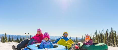 A group of people sitting on snow tubes on a snowy slope with a clear sky and trees in the background.