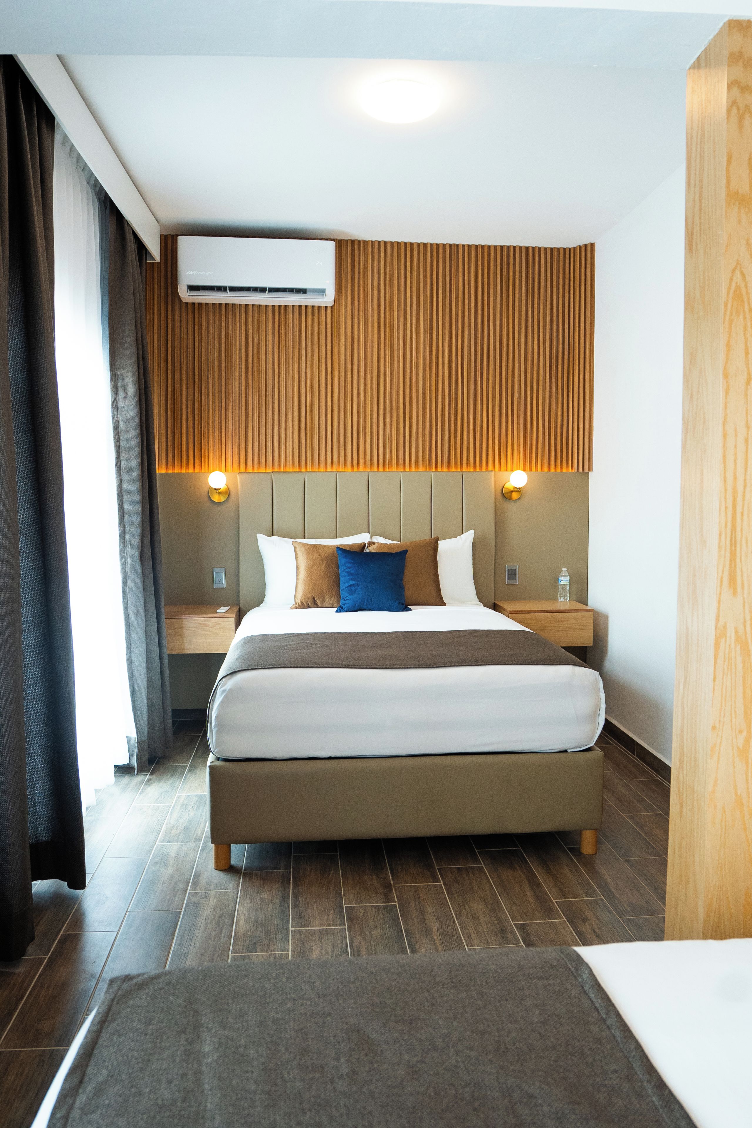 A hotel room with a bed, a nightstand, a wall-mounted air conditioner, and a wooden panel wall.
