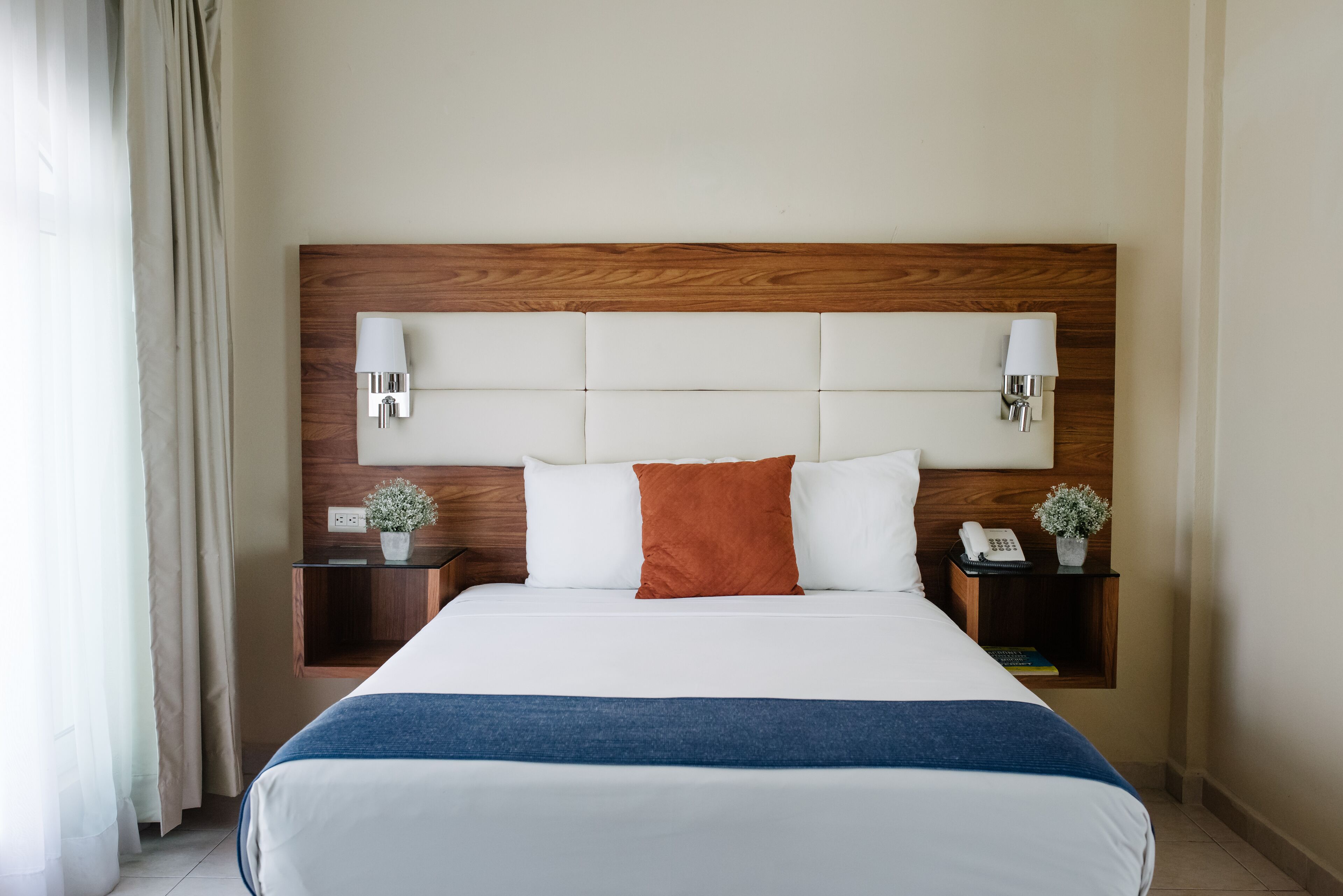Habitación de hotel con una cama grande, cabecera de madera, dos mesitas de noche con lámparas y un atril con teléfono y planta.