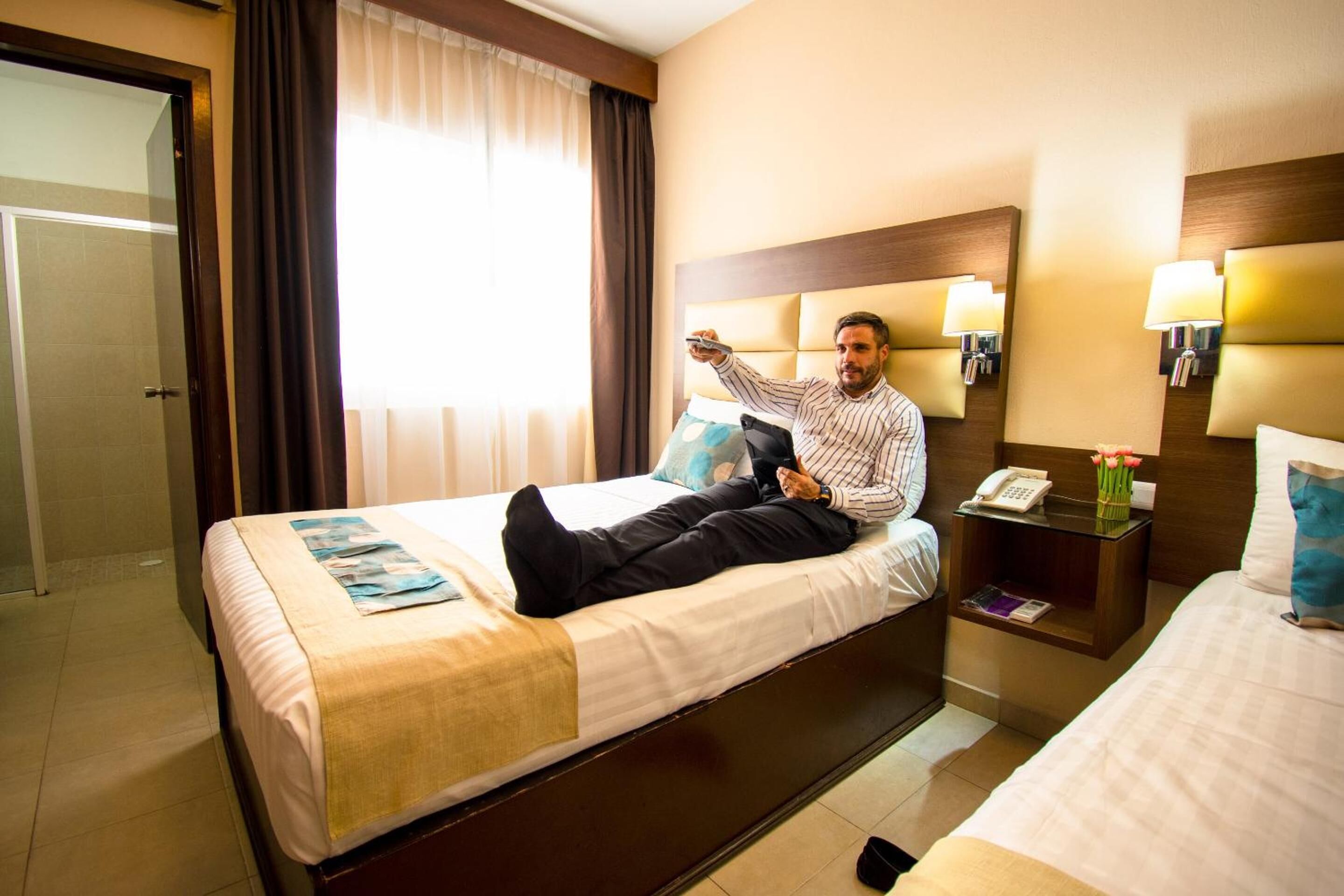 A hotel room with a bed, a bedside table with a lamp, a phone, and a nightstand with a vase of flowers.
