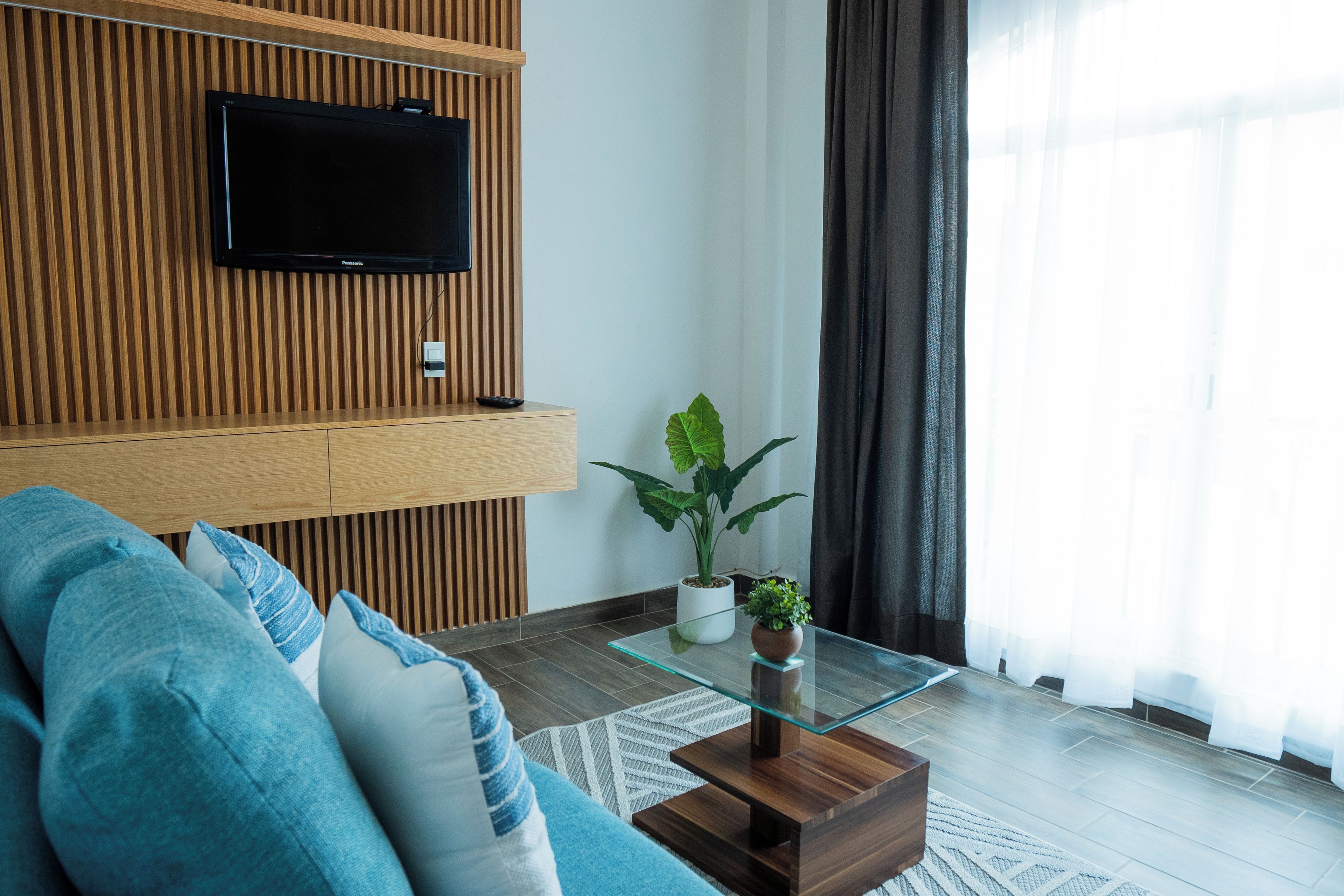 Una sala de estar moderna con un televisor de pantalla plana montado en una pared de madera, una mesa de centro de vidrio con una planta, y un sofá azul.