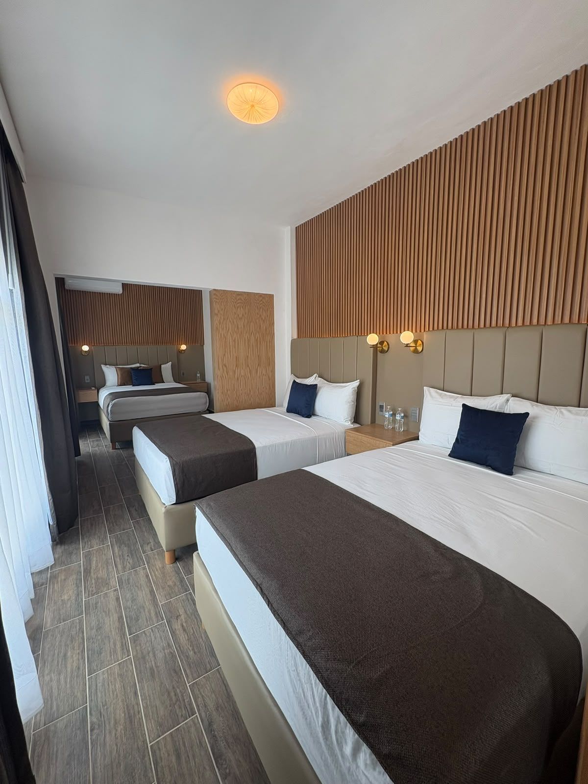 Habitación de hotel con dos camas, cabecero de madera, luz de techo y mesita de noche con lámpara.