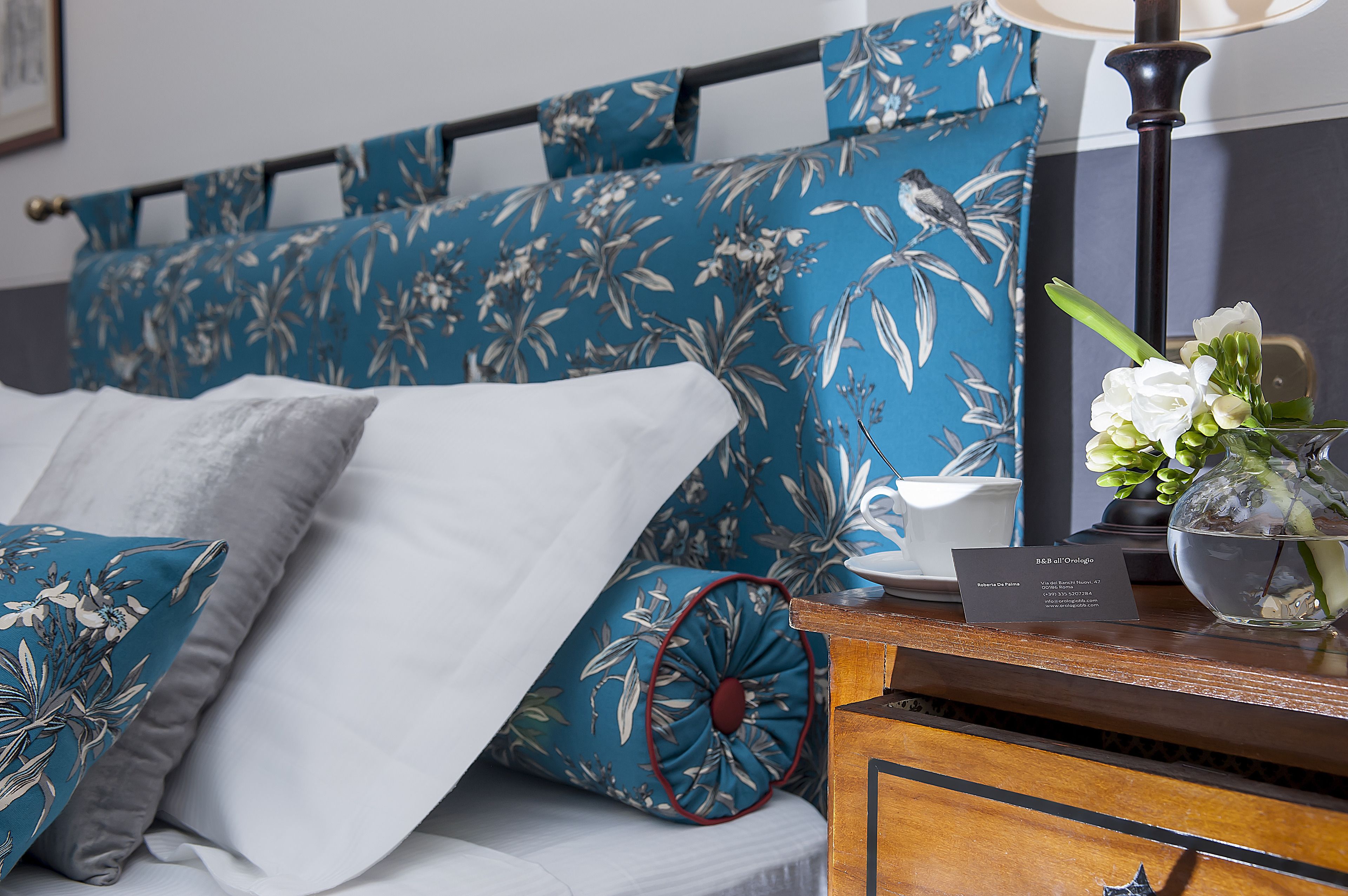 A neatly made bed with floral-patterned pillows and a bedside table with a lamp and a vase of flowers.