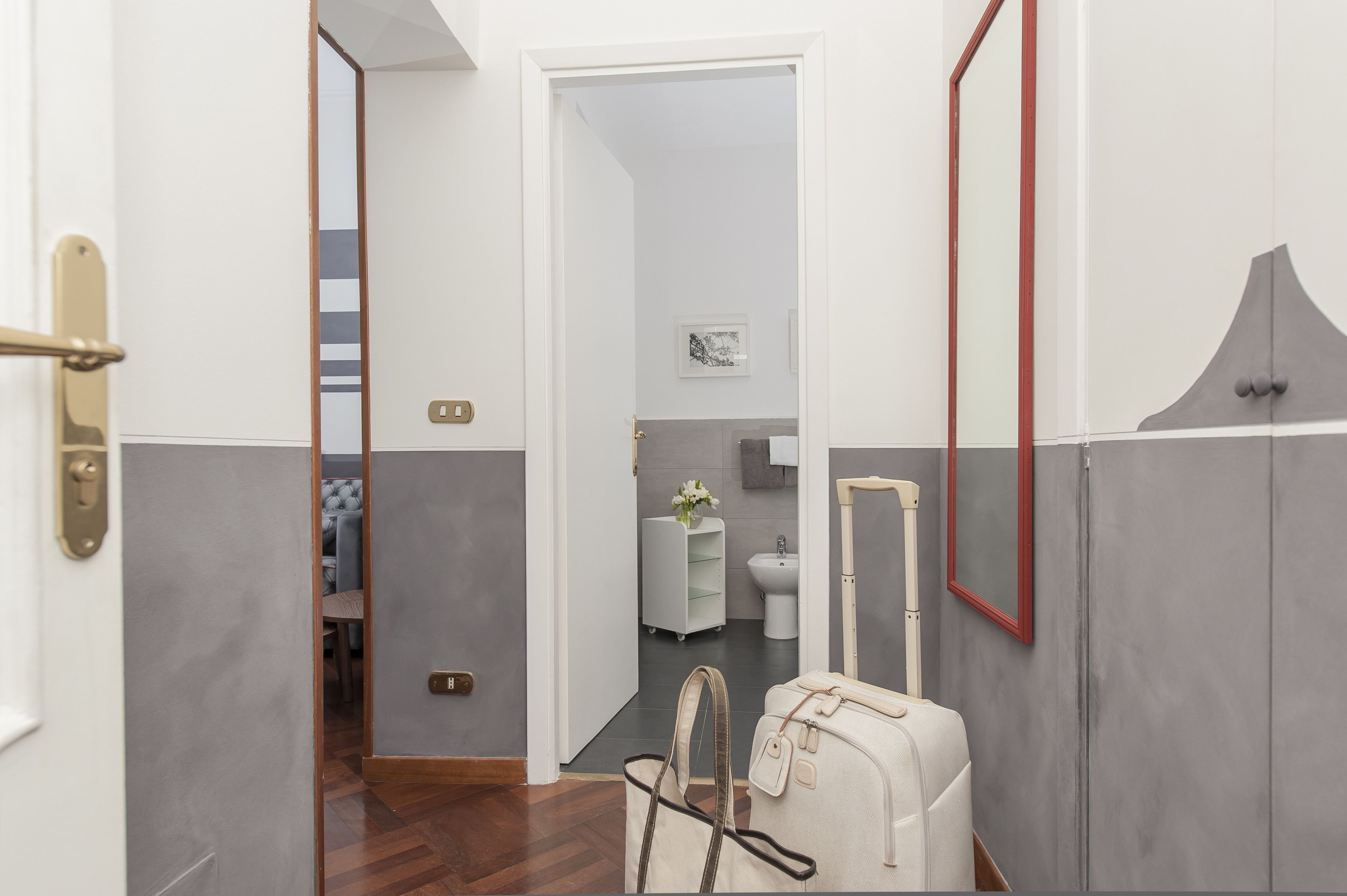 Um corredor com uma mala, uma porta e vista para um banheiro com vaso sanitário e pia.