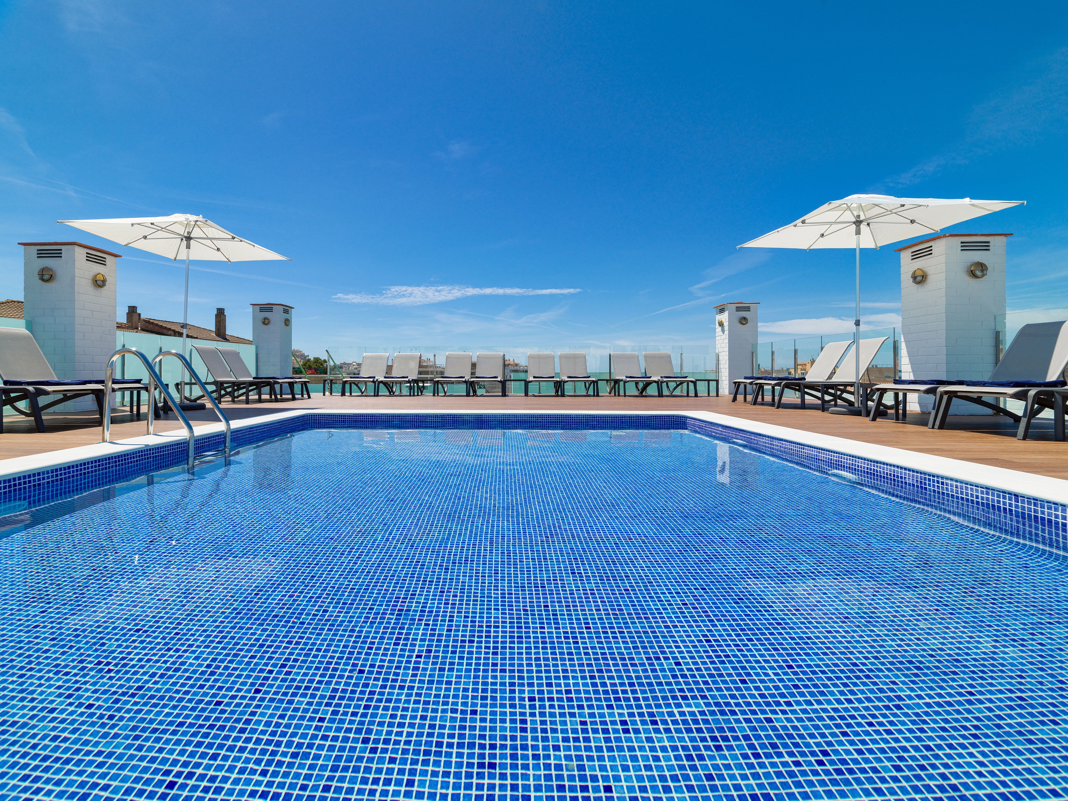 Una piscina con azulejos azules, rodeada de sillones de descanso y sombrillas, bajo un cielo despejado.