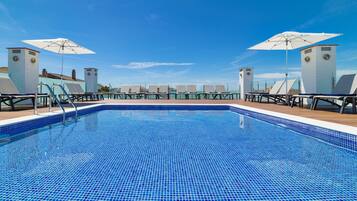 Una piscina con azulejos azules, rodeada de sillones de descanso y sombrillas, bajo un cielo despejado.