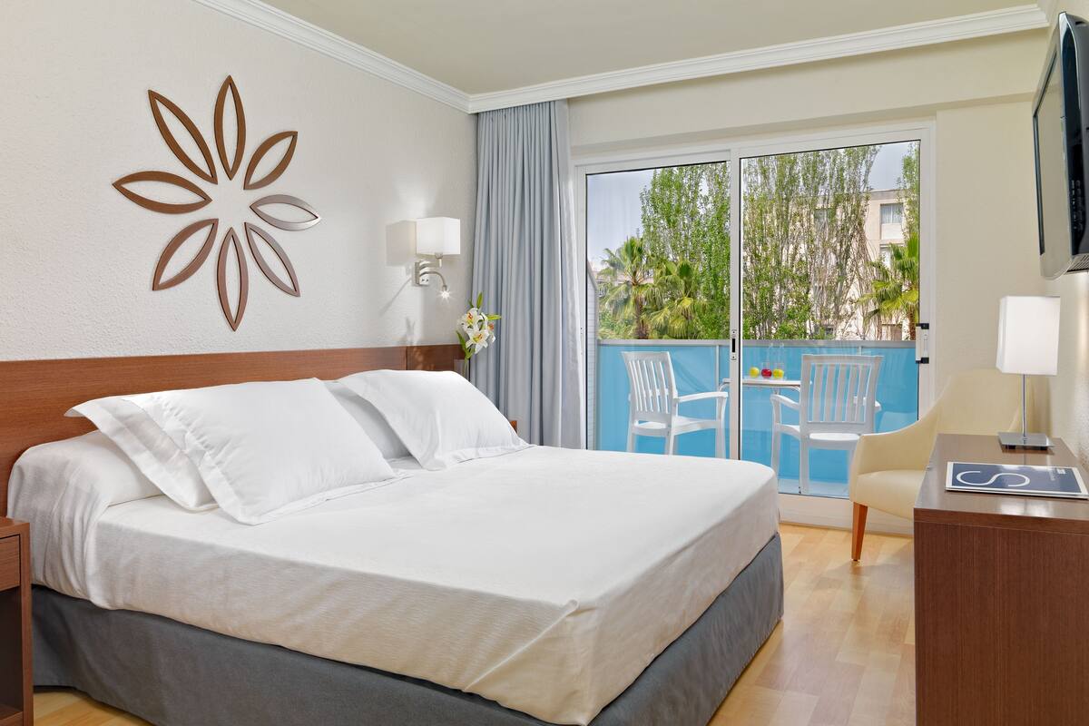 Habitación de hotel con una cama grande, un escritorio con lámpara, un florero y vistas al balcón exterior.