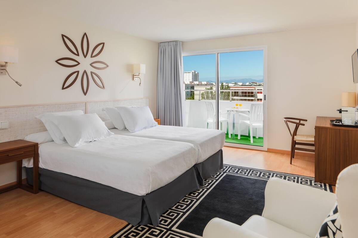 Habitación de hotel con dos camas, un escritorio, una silla y un balcón con vista a la ciudad.