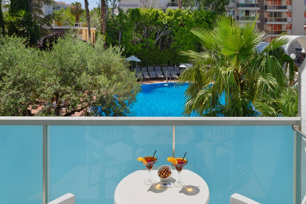 Un balcón con vista a la piscina, dos bebidas sobre una mesa y sillas blancas.