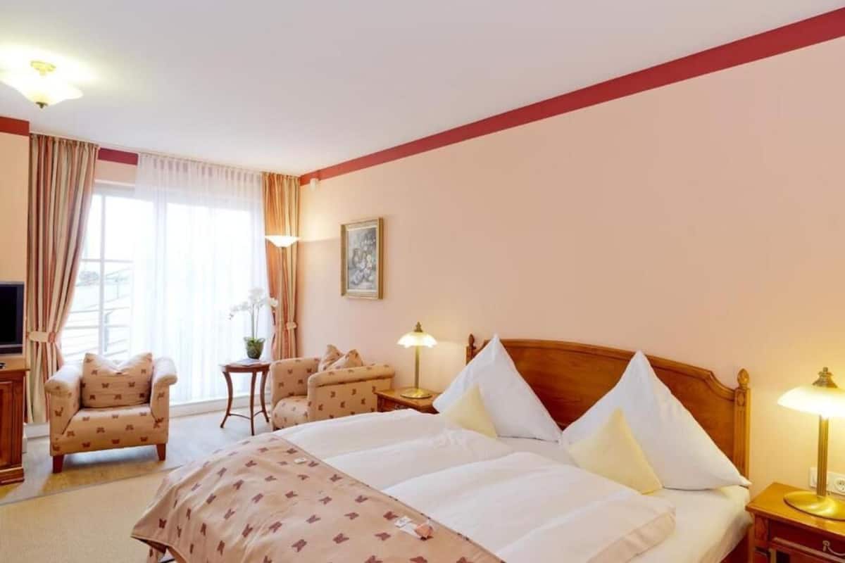 A hotel room with a large bed, two armchairs, a small table, a TV, and a window with curtains.
