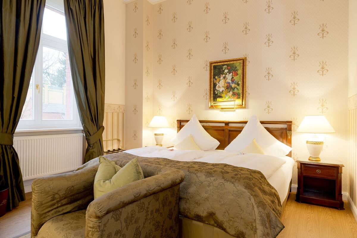 A hotel room with a large bed, two bedside lamps, a nightstand, a floral-patterned sofa, and a window with curtains.