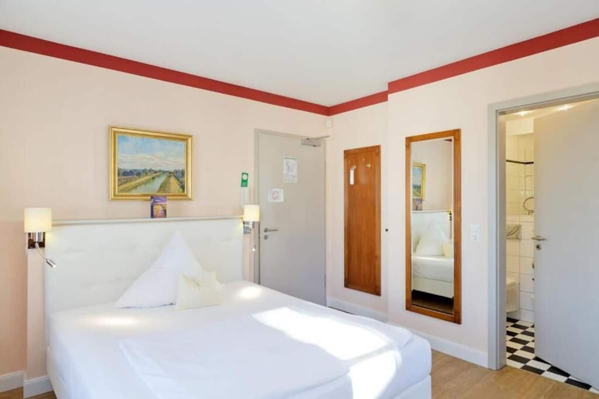 A hotel room with a bed, a painting on the wall, a door, and a mirror.
