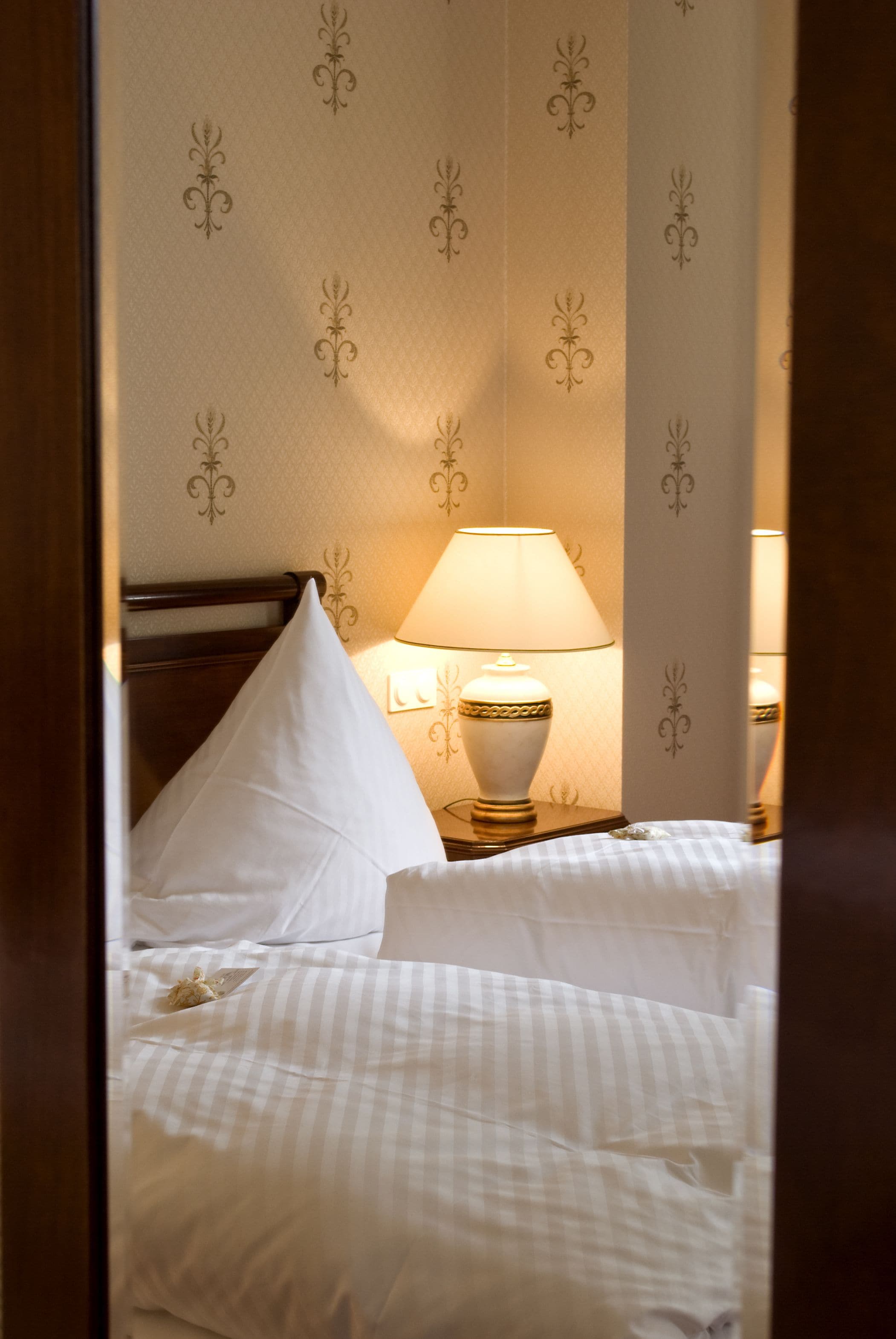 A neatly made bed with white linens and a striped pillow, a bedside lamp with a decorative base, and a wall with a repeating floral pattern.