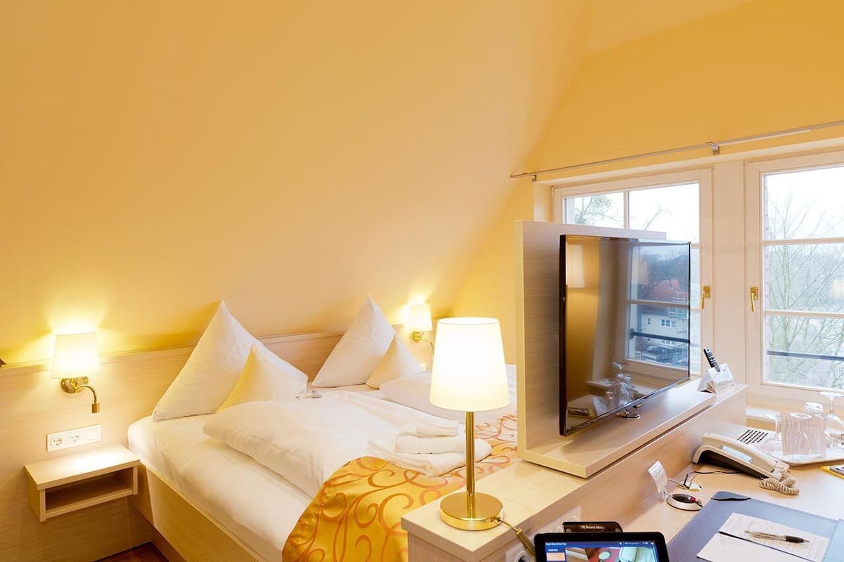 A hotel room with a bed, bedside lamps, a desk with a phone, a mirror, and a window with a view of buildings.