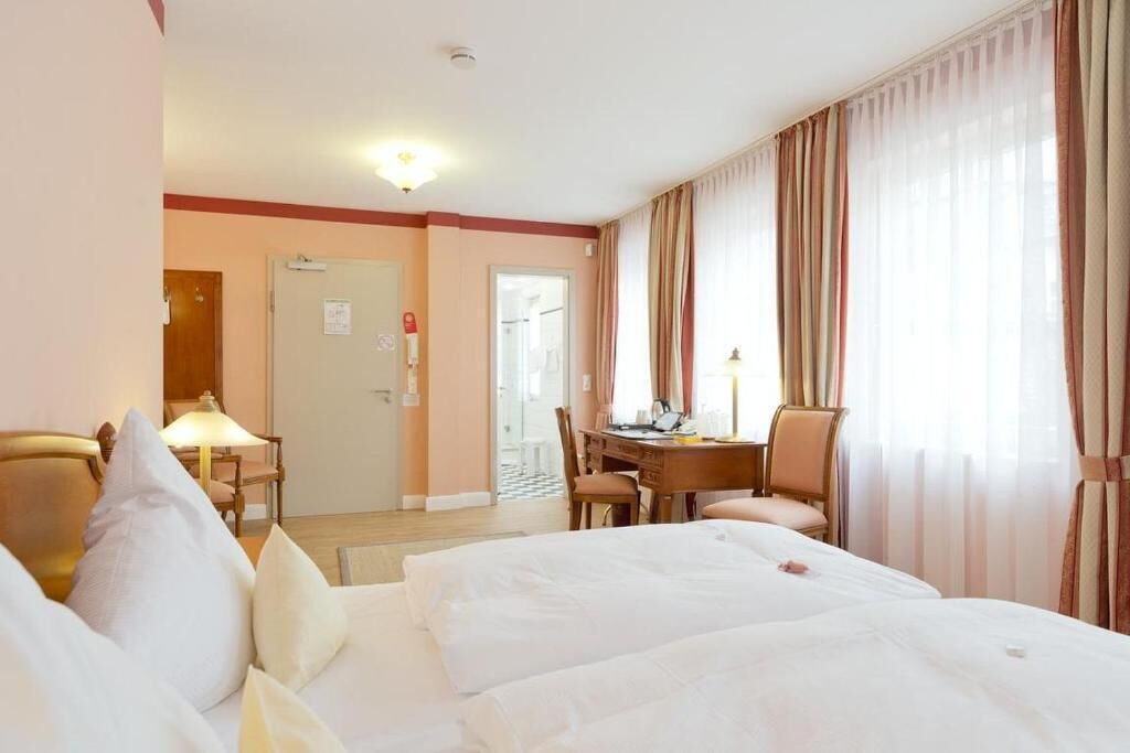 Une chambre d’hôtel avec un lit, un bureau et une chaise. Il y a une porte qui donne sur une autre pièce et une fenêtre avec des rideaux.