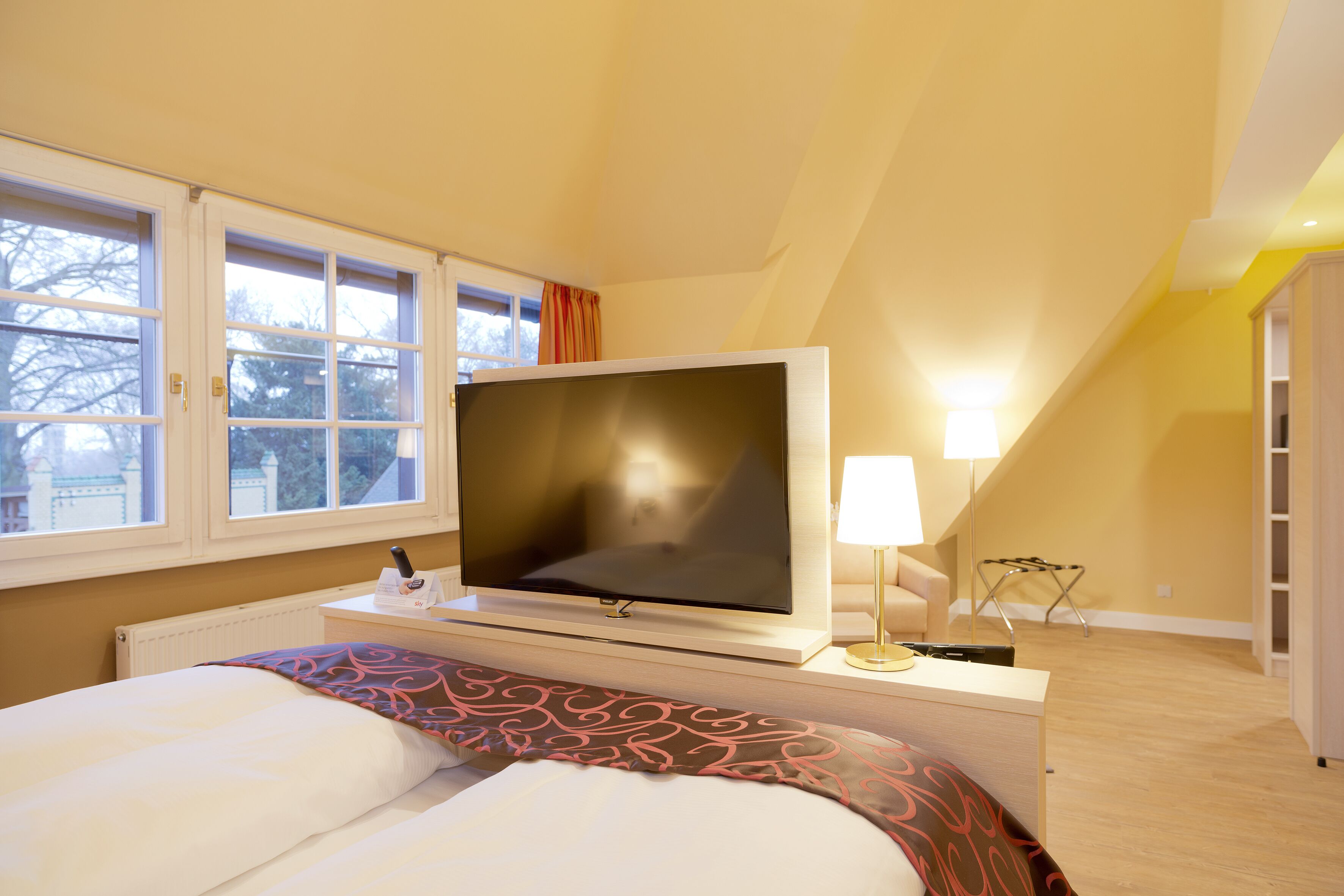 A hotel room with a bed, a flat-screen TV, a bedside table with a lamp, and a window with curtains.