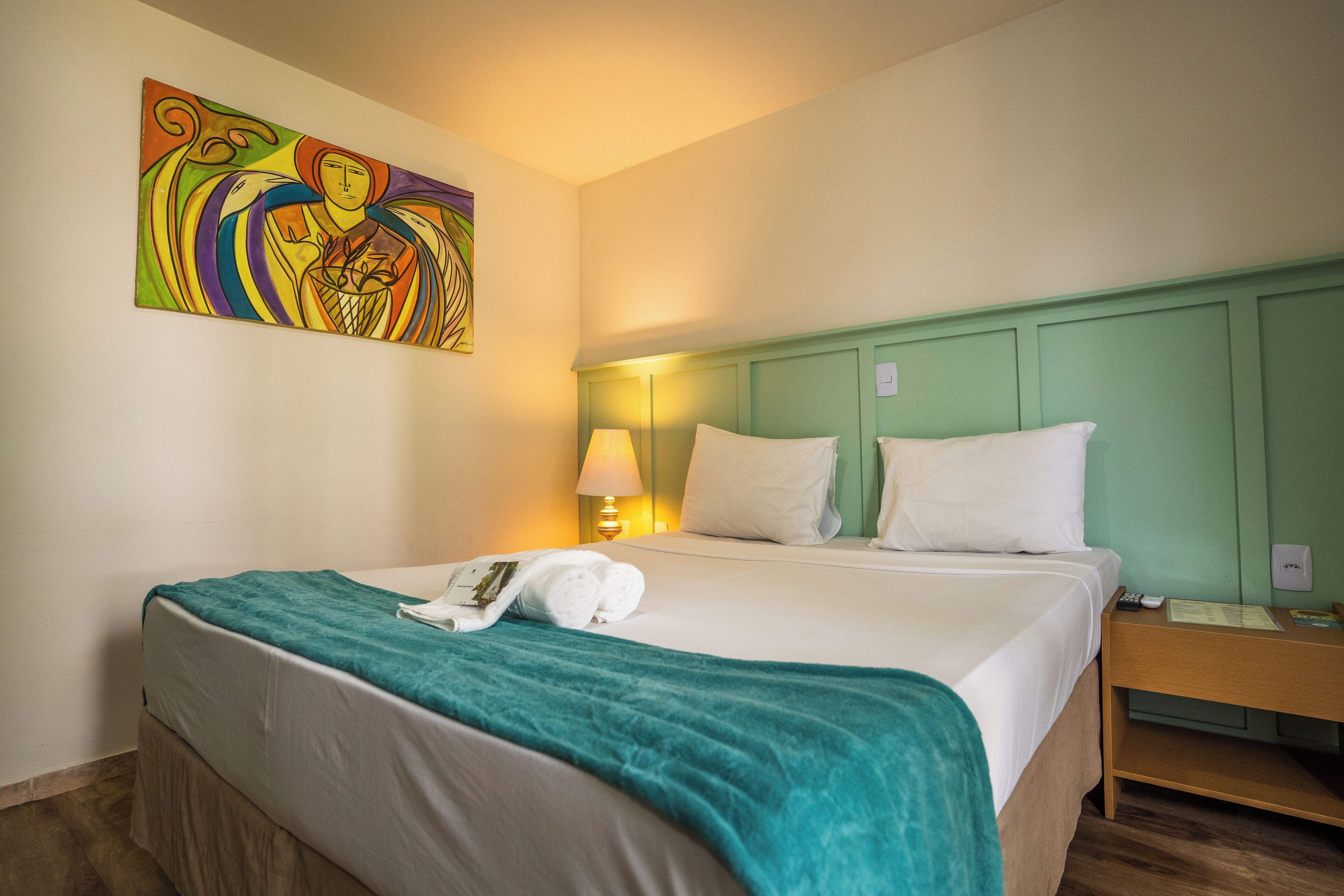 Una habitación de hotel con cama, una mesita de noche con lámpara, un cuadro en la pared y una manta azul turquesa sobre la cama.