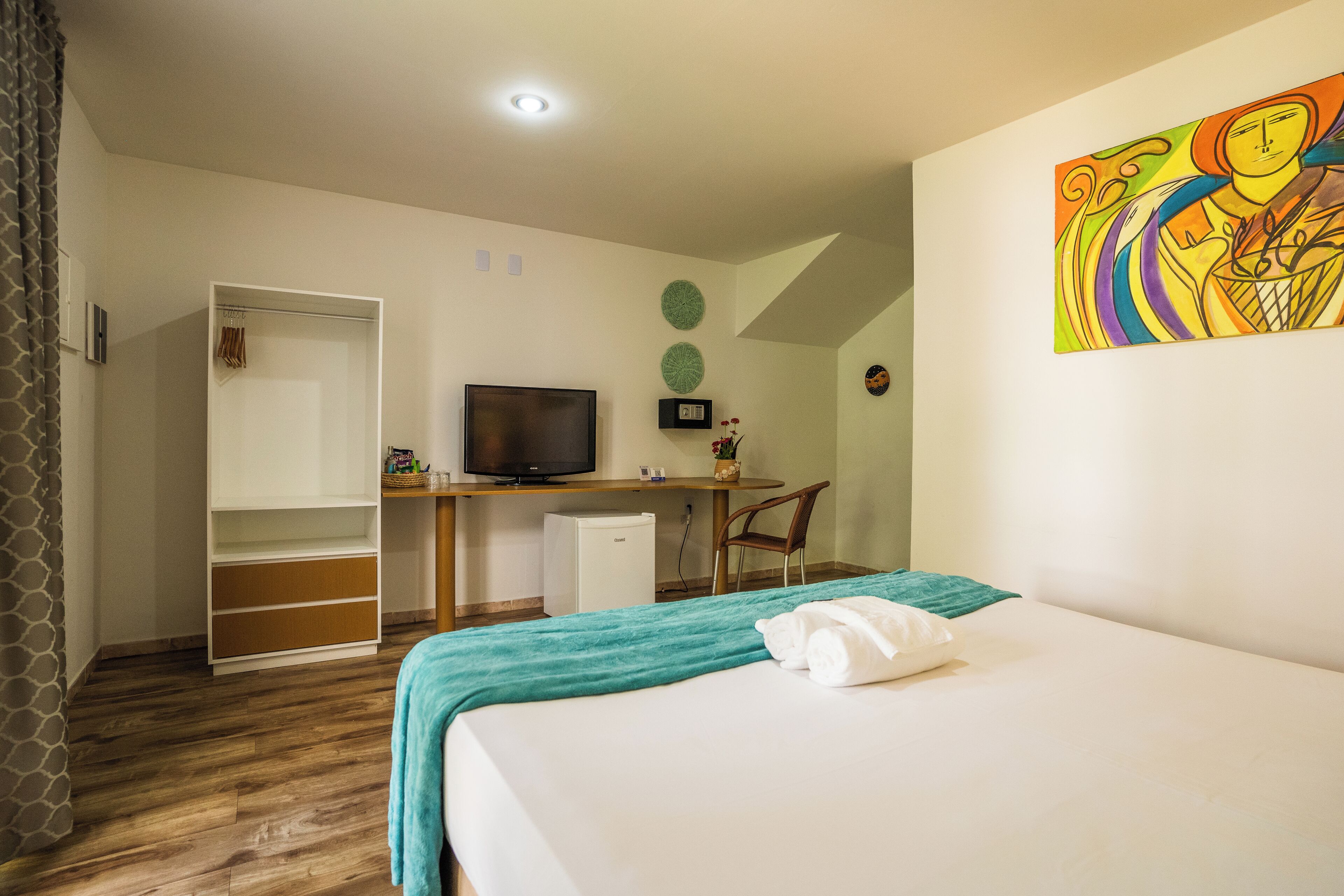A hotel room with a bed, a desk with a television, a wardrobe, and a small refrigerator.
