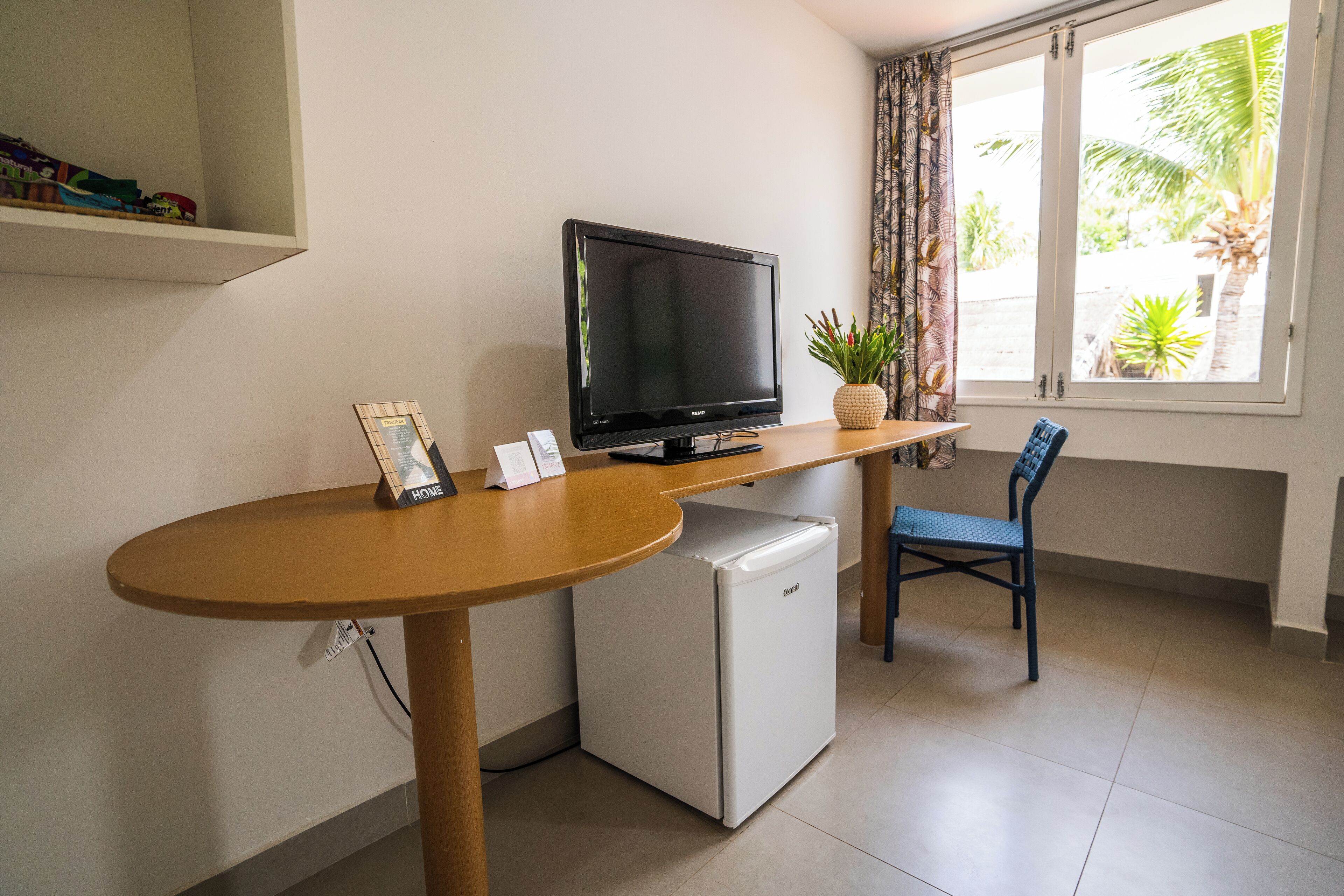 Un espace de vie compact comprenant une télévision, un petit réfrigérateur, une table à manger, une chaise et une fenêtre donnant sur des palmiers.