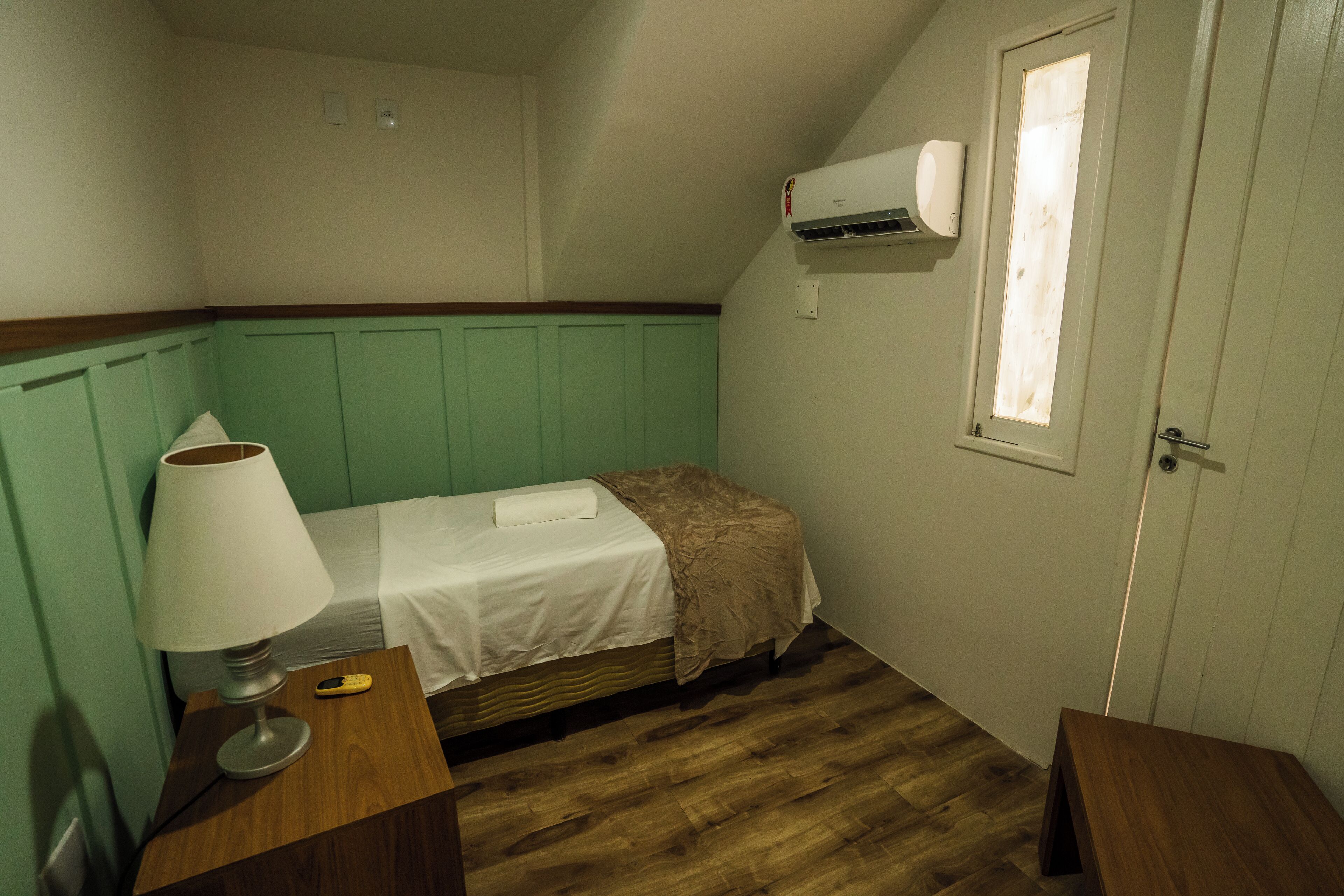 A small, single-bed room with a wooden floor, a bedside table with a lamp, and a window with a view of the outside.