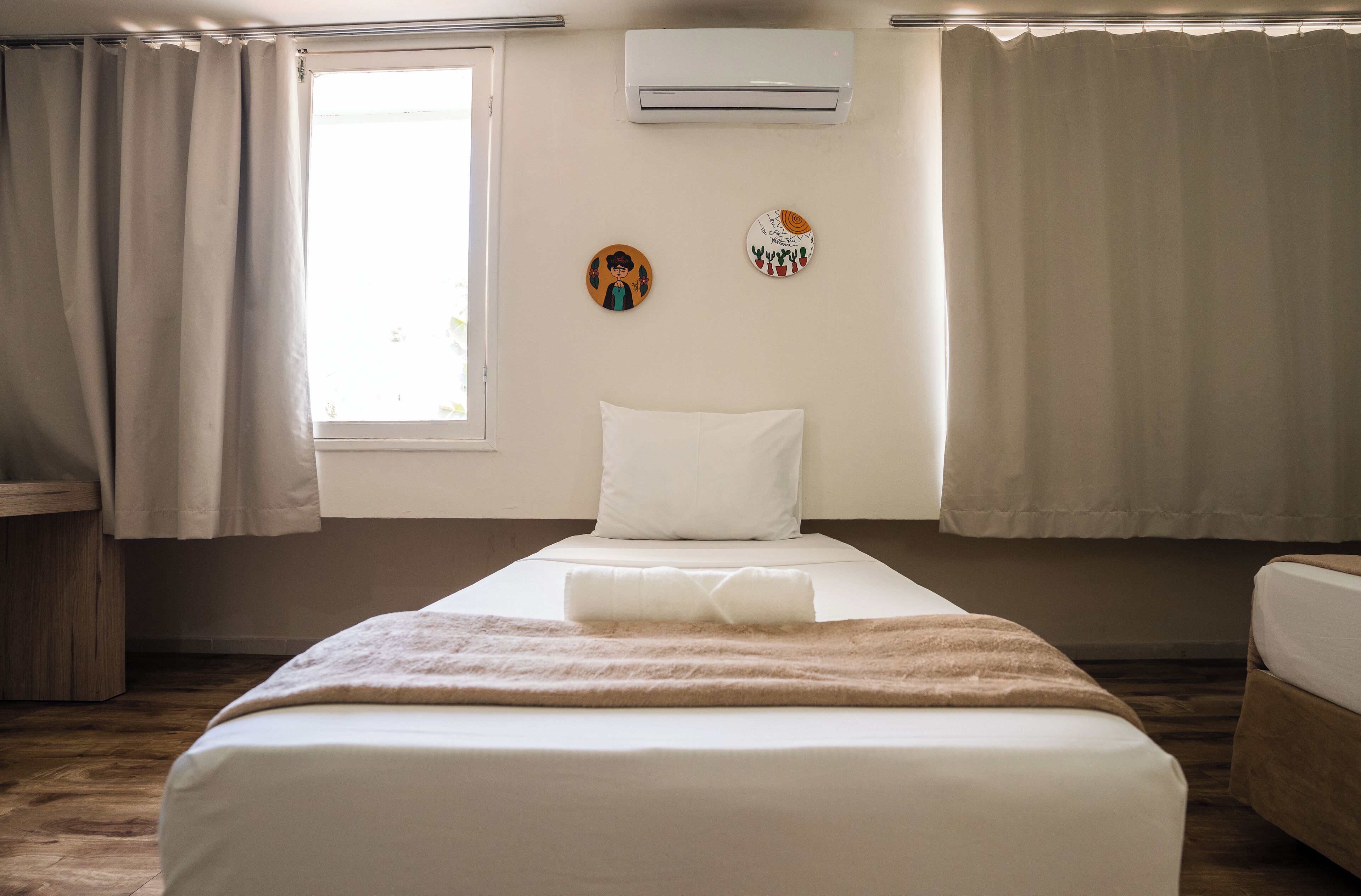 Uma cama de solteiro com travesseiro branco e manta marrom, uma janela com cortinas e duas decorações na parede.