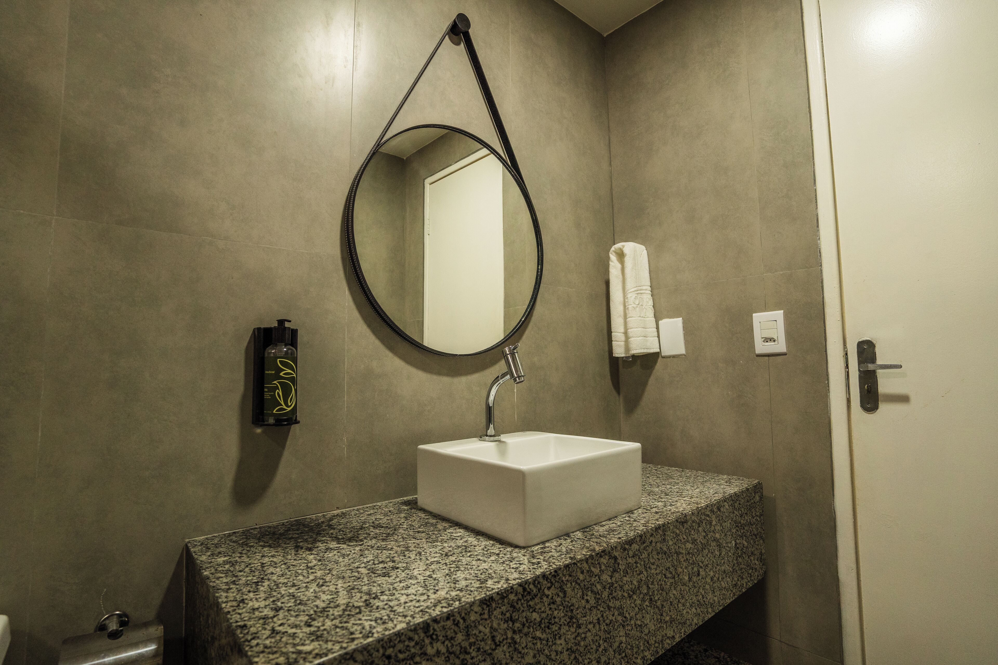 A bathroom with a granite countertop, a white sink, a round mirror, and a towel dispenser.