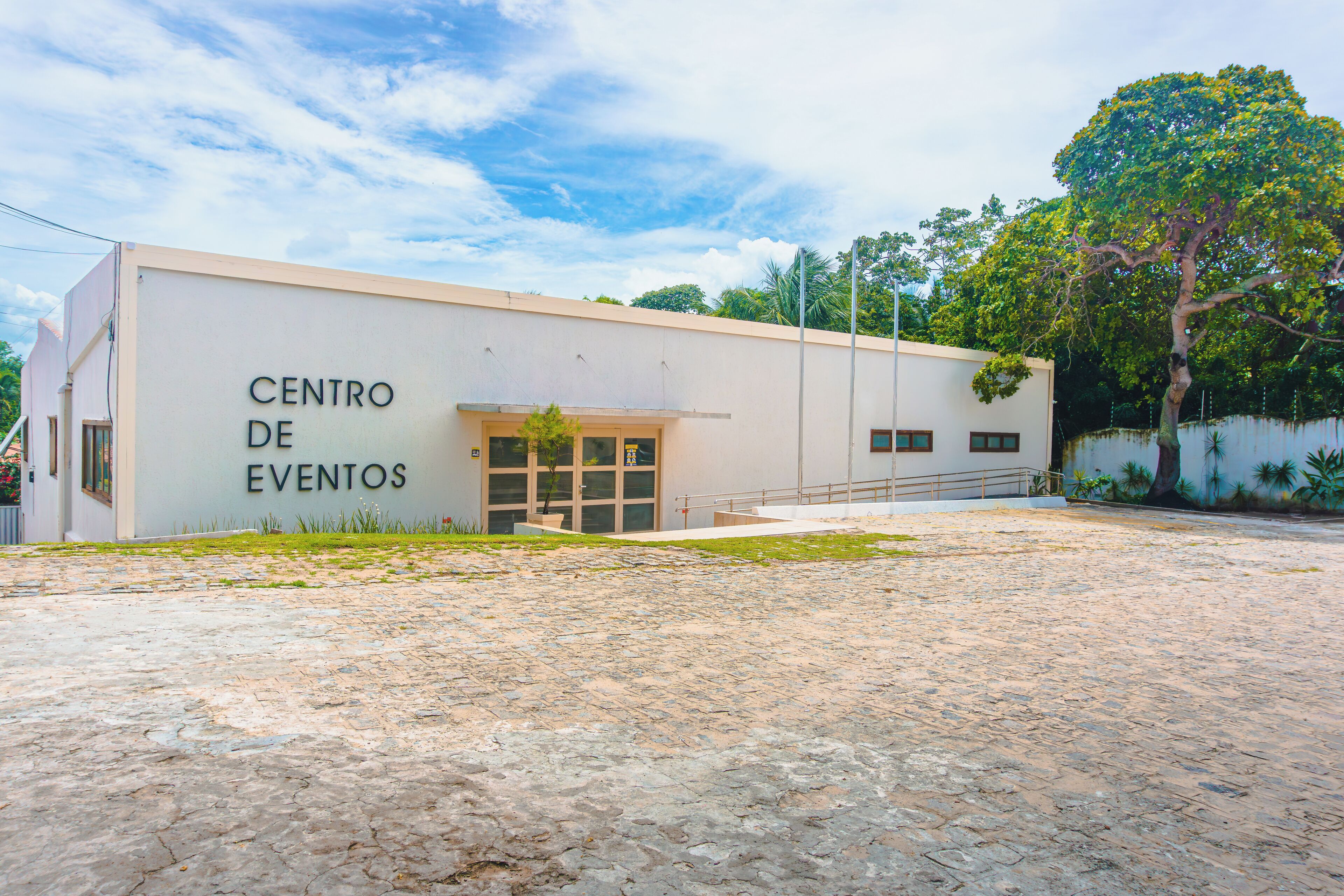 Un edificio con la leyenda "CENTRO DE EVENTOS", un área de acceso pavimentada y un árbol cercano.