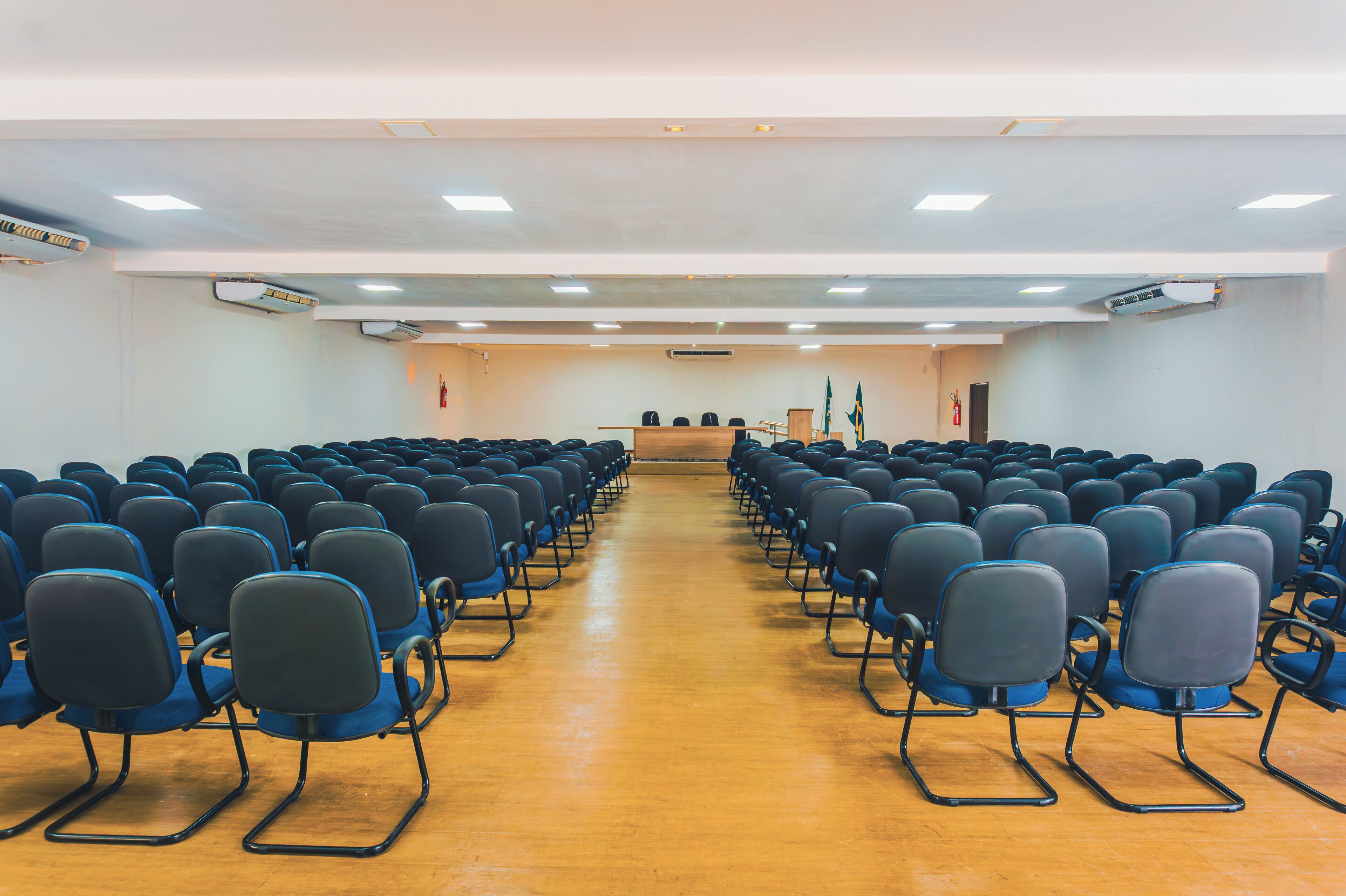 A conference room with rows of chairs facing a podium, equipped with an air conditioning unit and flags.