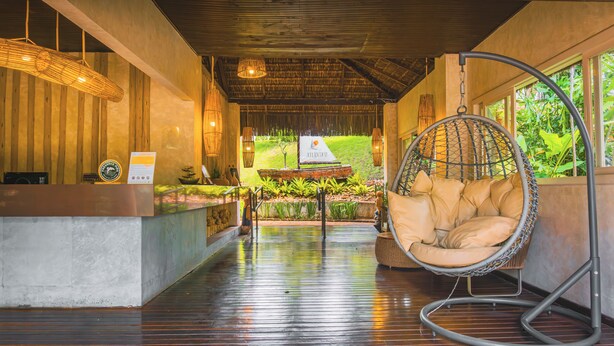 Zona de recepción con piso de madera, mostrador de recepción, una silla colgante y vista a un jardín a través de amplias ventanas.