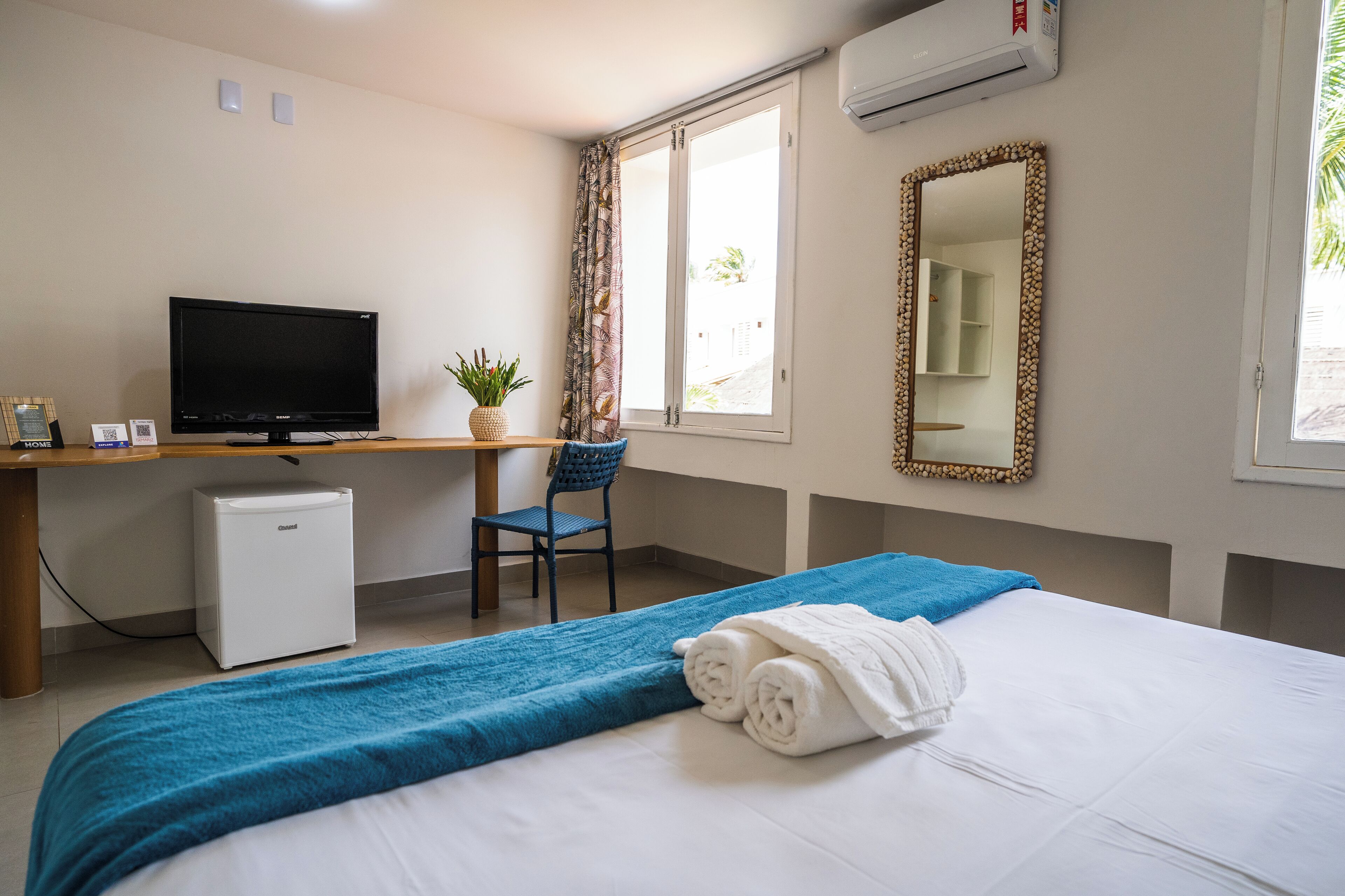 A hotel room with a bed, a desk with a television, a small refrigerator, a chair, and a window with curtains.
