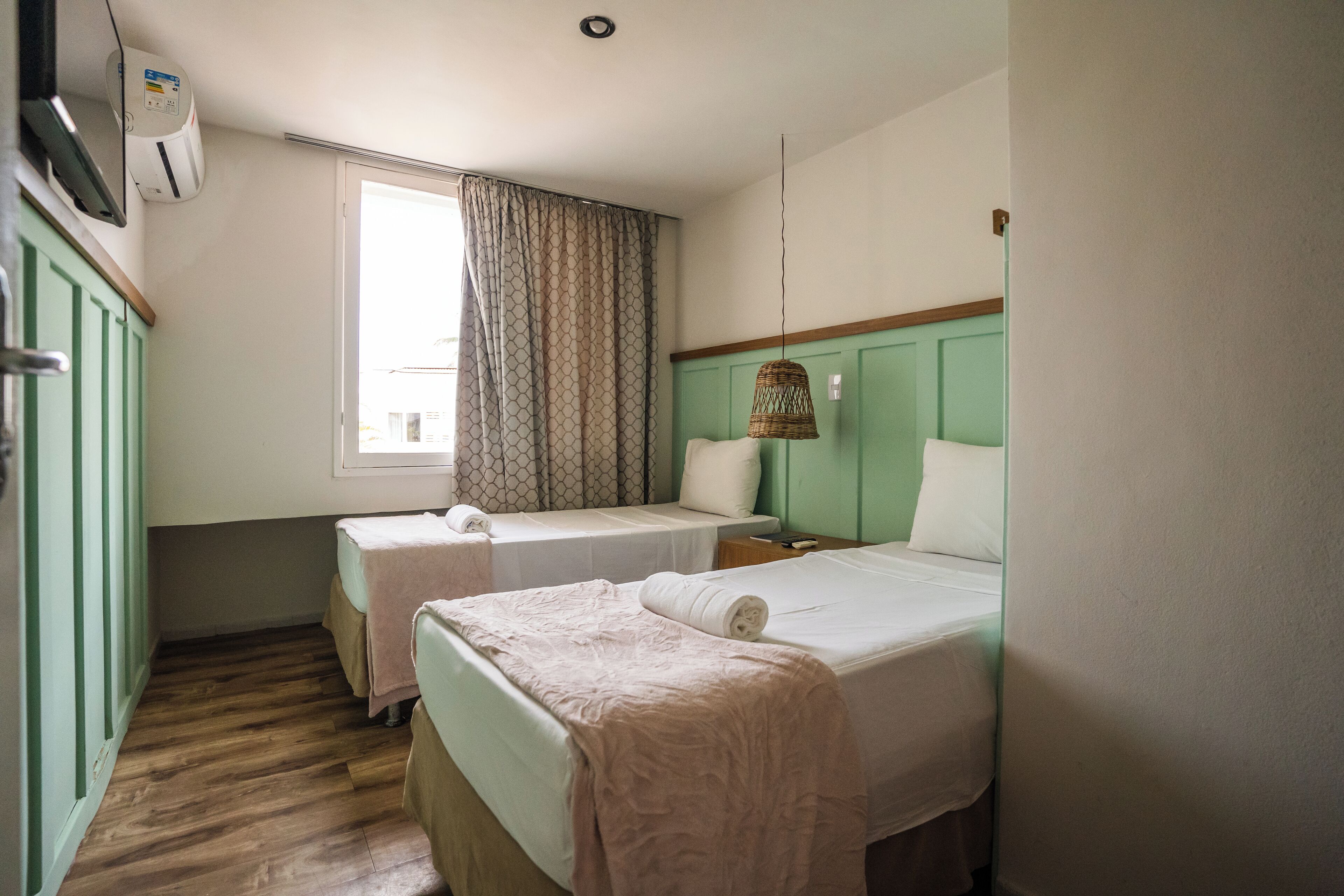 Una habitación con dos camas, una ventana con cortinas, un aire acondicionado montado en la pared y una lámpara colgante.