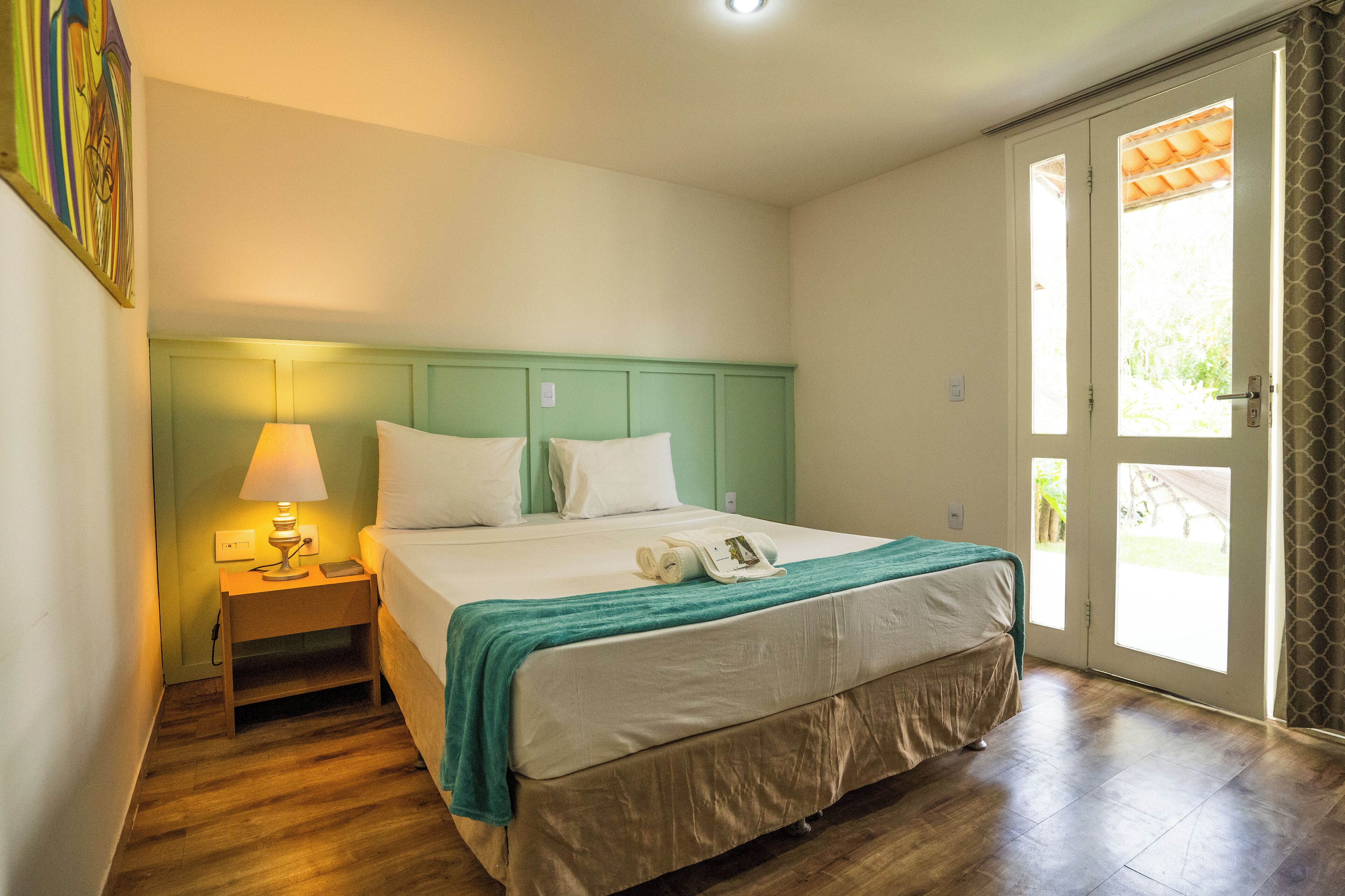 Quarto de hotel com uma cama, uma mesa de cabeceira com abajur e vista para o exterior através de uma porta.