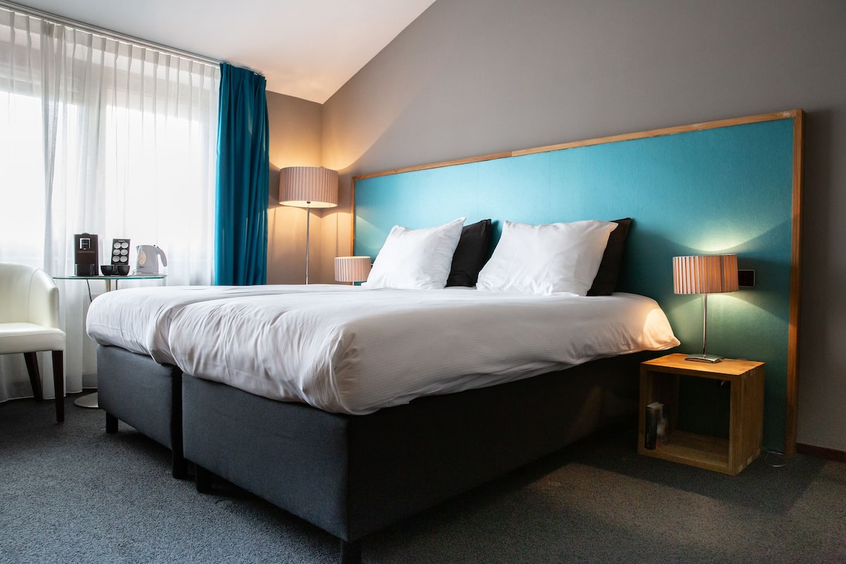 A modern hotel room with a large bed, a desk with a chair, a coffee maker, and a lamp.