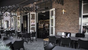 An outdoor cafe with tables and chairs, open doors leading to the interior, and a brick wall. - Linge Hotel Elst (Elst)