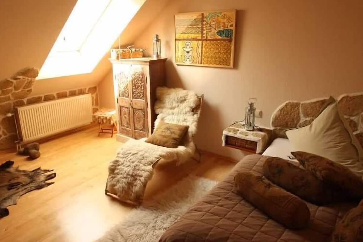 Ein gemütliches Schlafzimmer im Dachgeschoss mit schrägen Wänden, Steinwand, Holzboden, einem Bett mit Felldecke, einem kleinen Holzstuhl und einer Heizung.