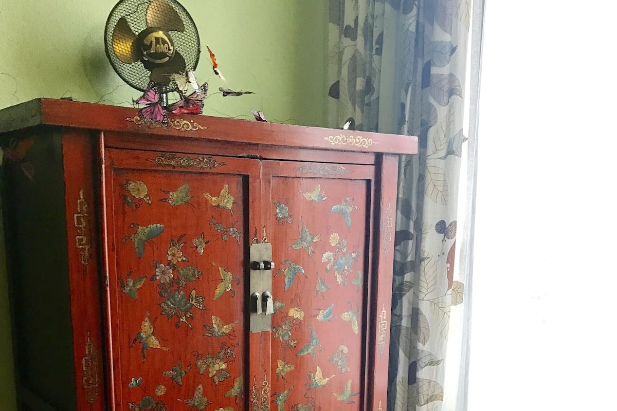 Ein roter Korpus mit floralen und Vogelmotiven, ein kleiner Fächer obenauf und ein Stuhl mit einem Kissen in Blätermuster.