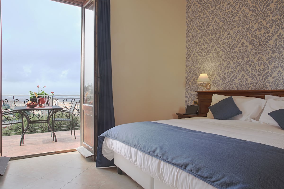 A hotel room with a bed, a nightstand, a lamp, a table with chairs, and a view of the city through a balcony.