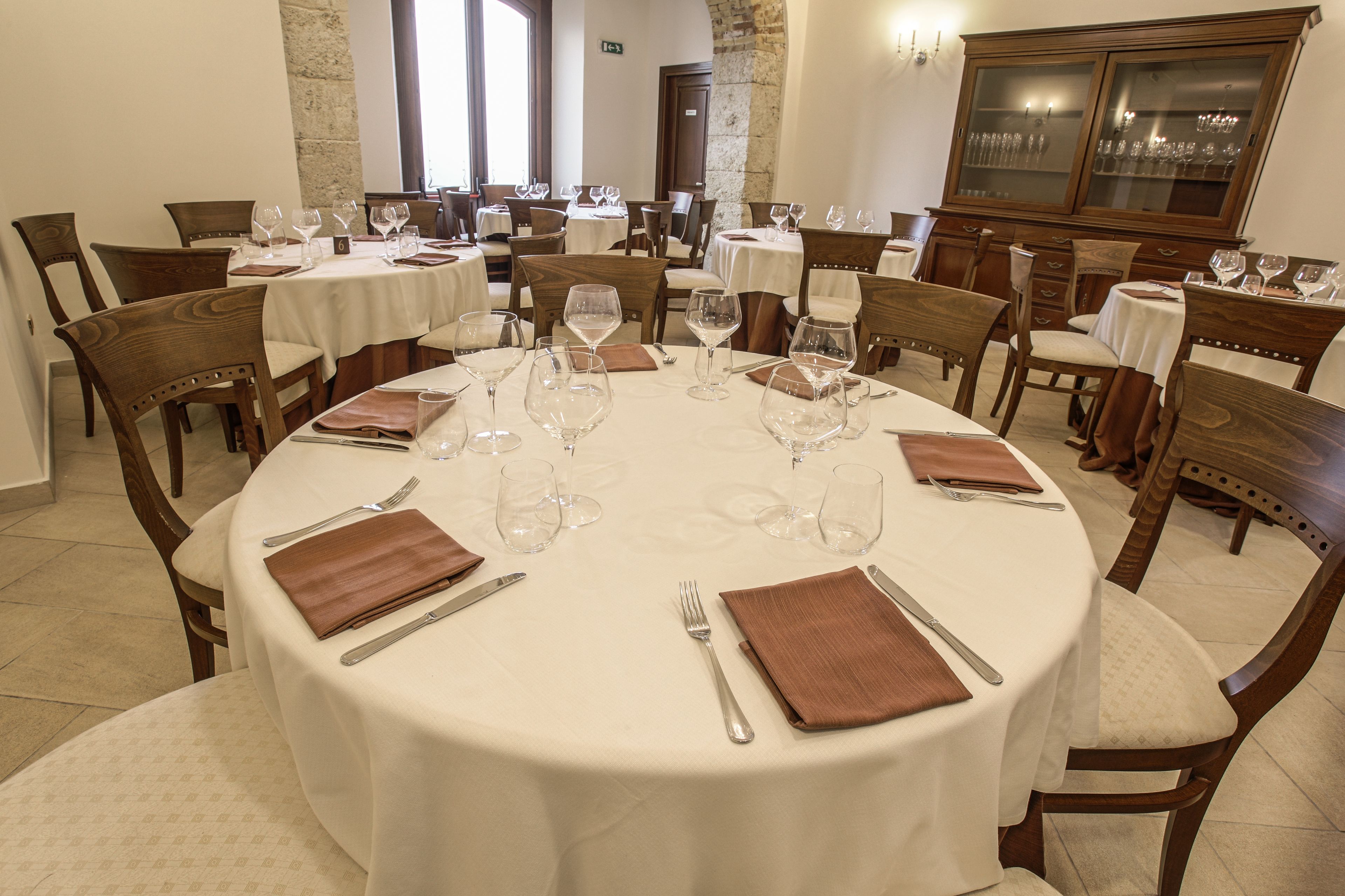 A dining area with round tables set for a meal, each with a white tablecloth, brown napkins, and cutlery.
