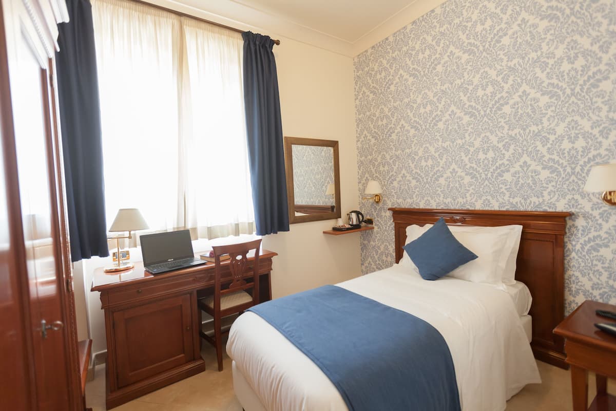 A hotel room with a bed, a desk with a laptop, a chair, a nightstand, and a window with curtains.