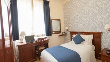 A hotel room with a bed, a desk with a laptop, a chair, a nightstand, and a window with curtains.