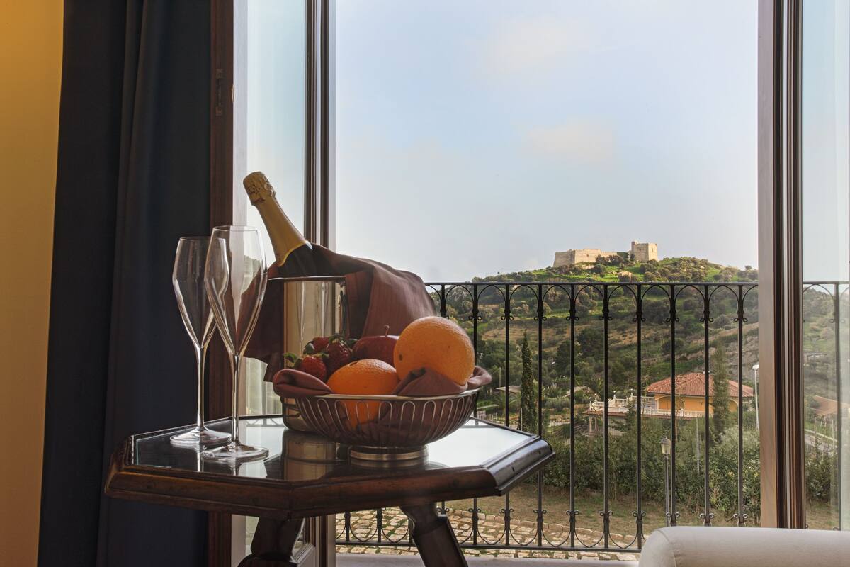 A table with a fruit bowl, a champagne bottle, and two glasses on a balcony overlooking a scenic view of a hill with buildings.