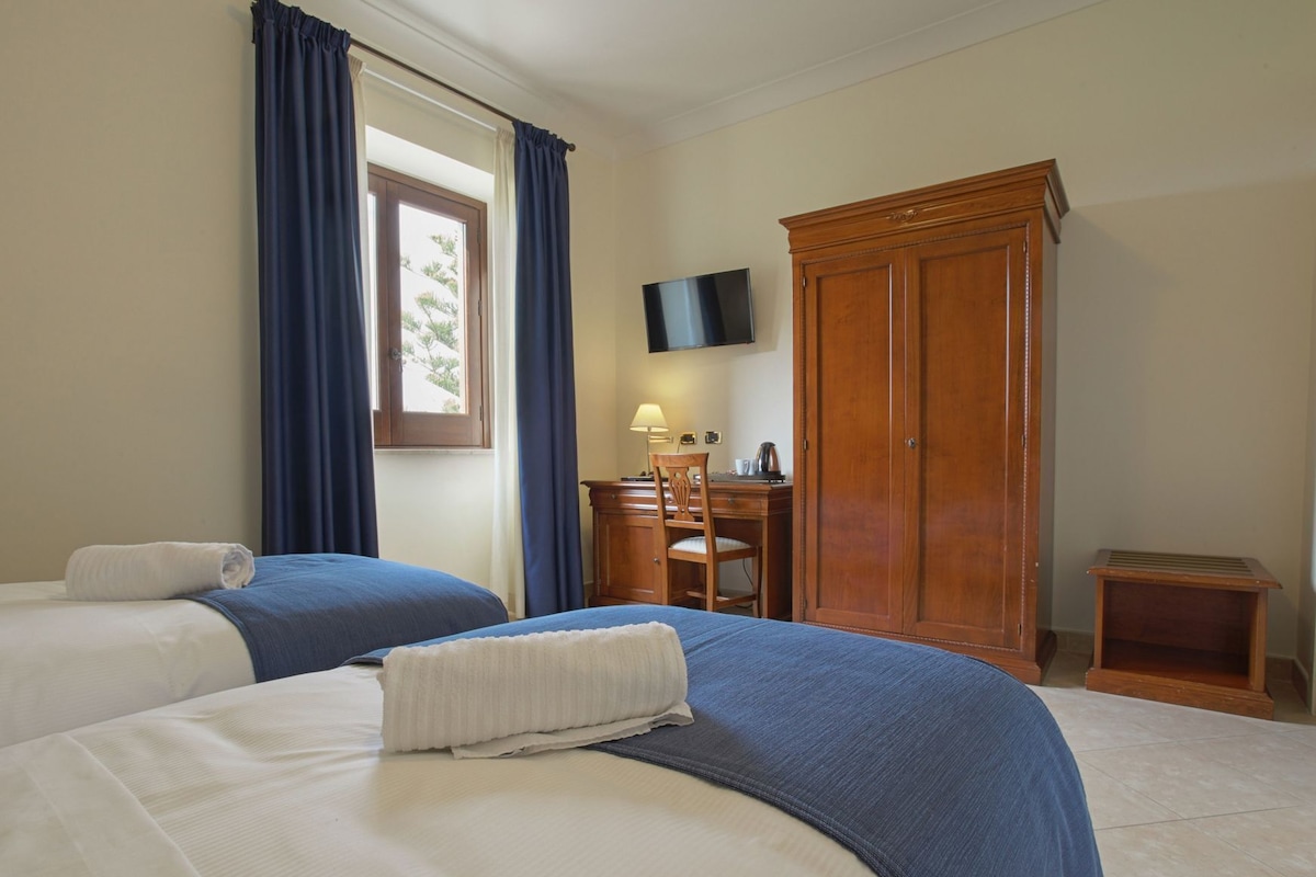 A hotel room with two beds, a wooden wardrobe, a desk with a chair, a television, and a window with curtains.